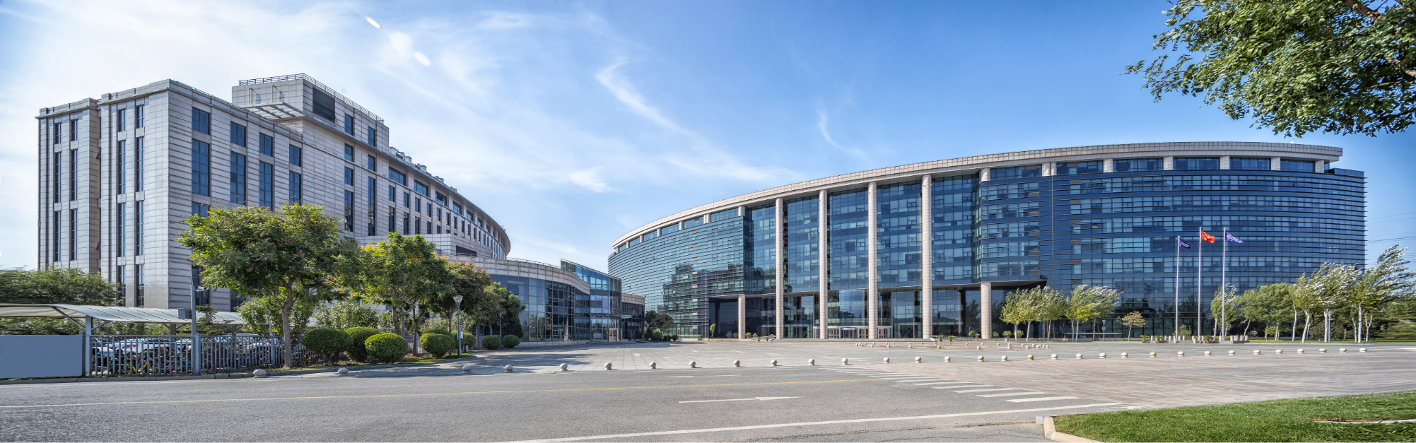 Two modern curved glass office buildings with trees and flags in front, under a blue sky.