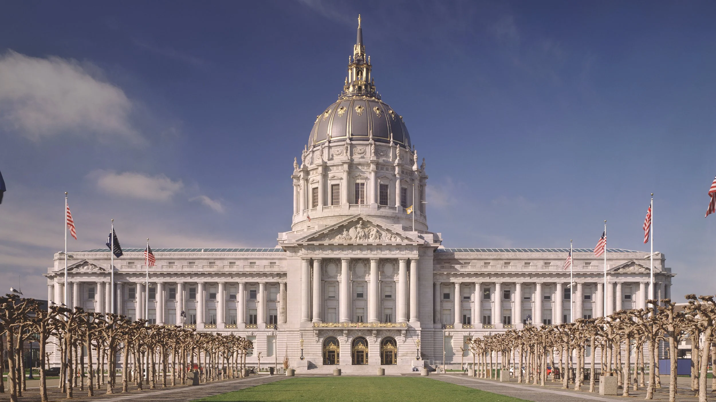 The California State Capitol building in Sacramento, with a large central dome, surrounded by smaller structures, American flags, and pruned trees in the foreground.