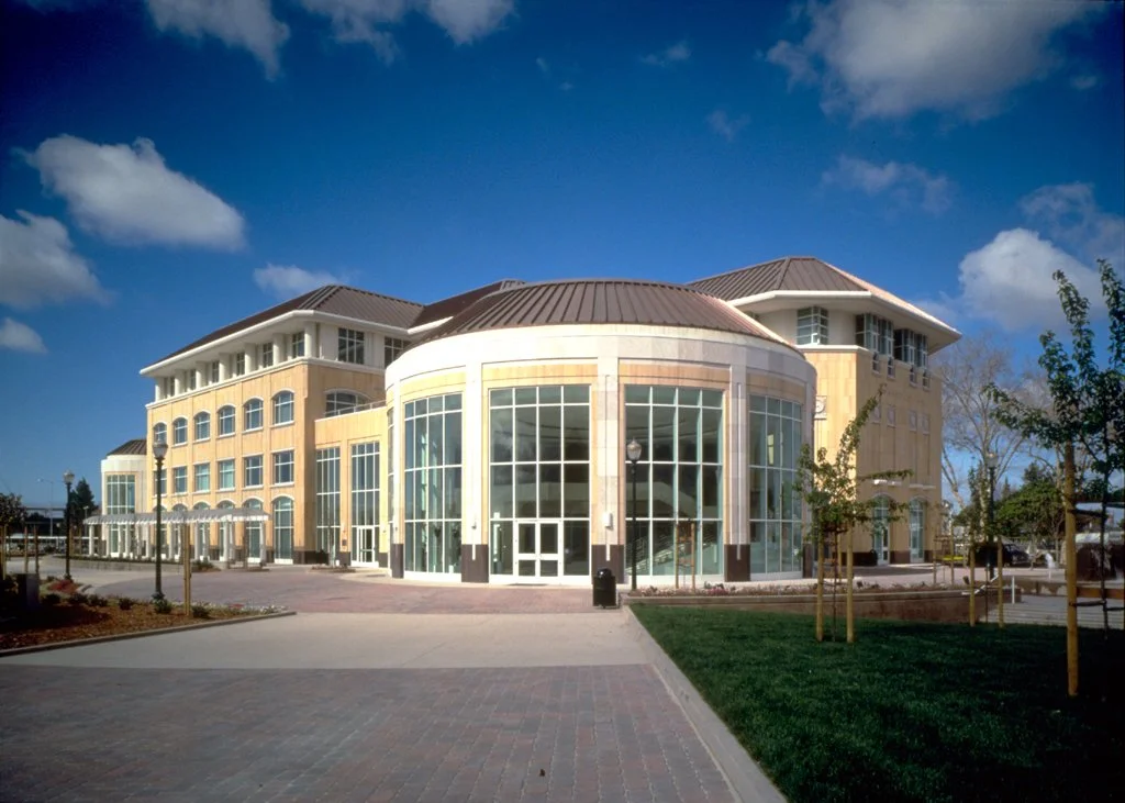Modern multi-story building with large glass windows and a rounded entrance, surrounded by a paved plaza and small trees, under a blue sky with some clouds.