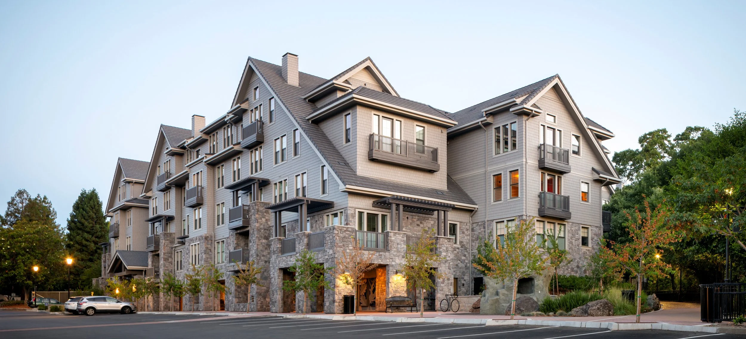Multi-story residential building with stone and wood exterior, multiple balconies, surrounded by trees, parking lot with cars, and streetlights at dusk.