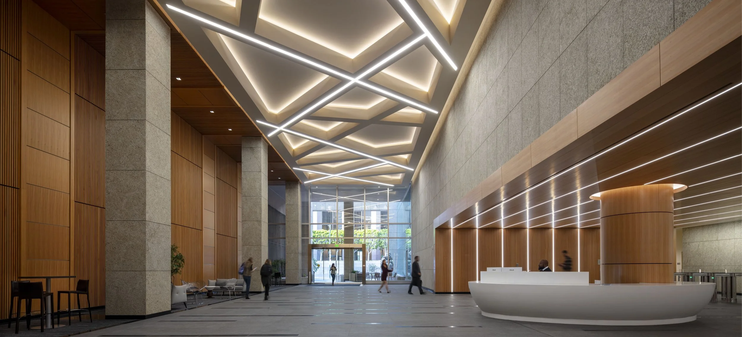 Modern hotel lobby with a high ceiling featuring geometric lighting fixtures, wooden and stone wall accents, a reception desk with a receptionist, and a few people walking or sitting.