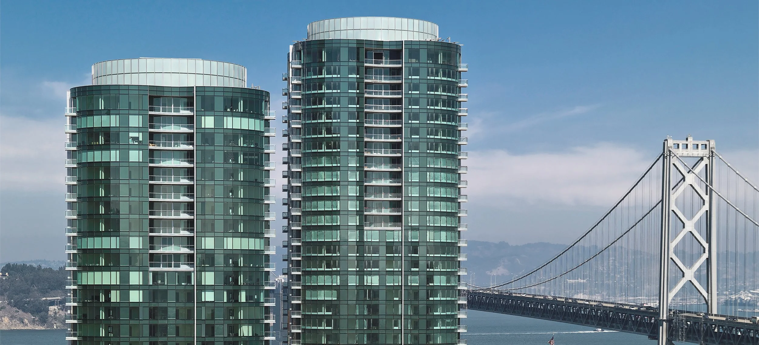 Two modern green glass high-rise buildings with a bridge in the background and a blue sky.