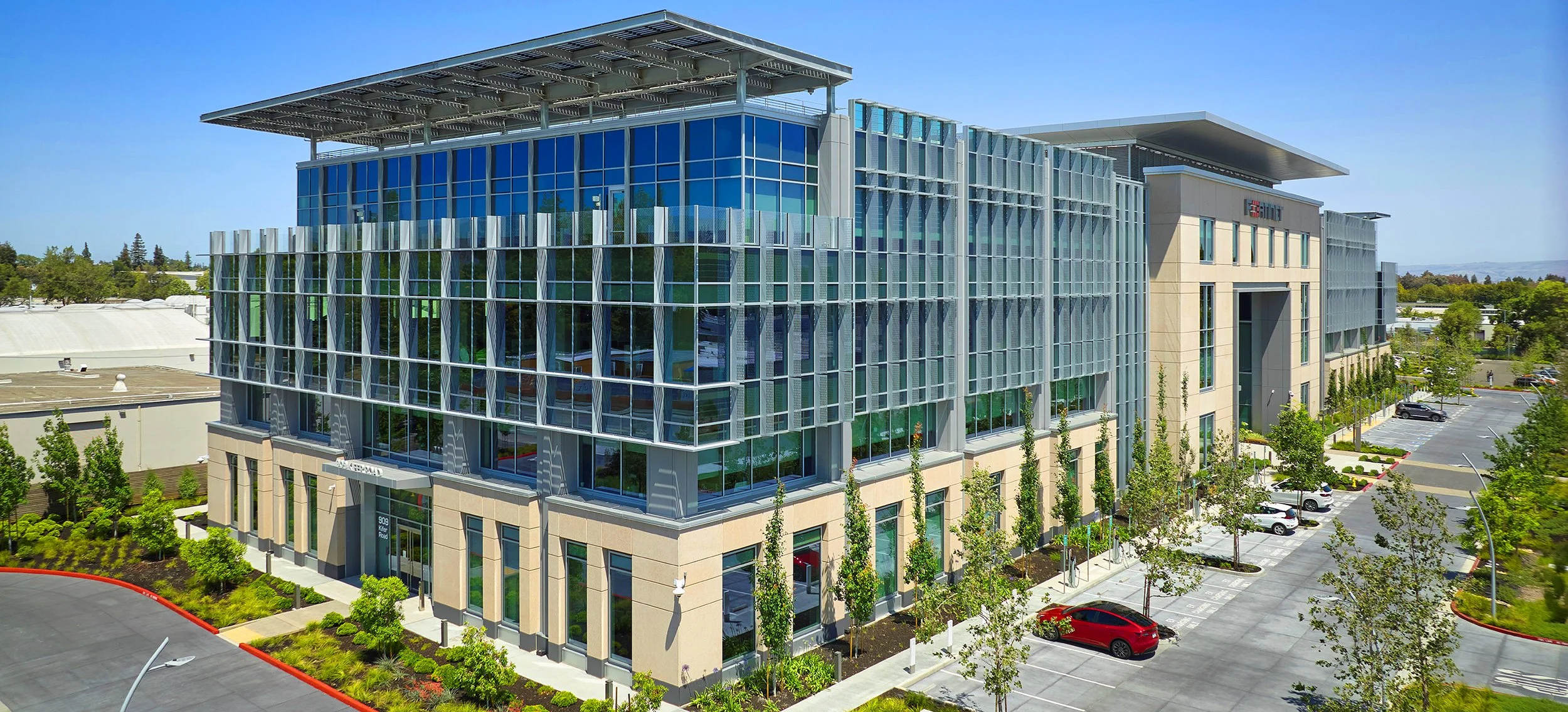 Modern office building with glass windows, surrounded by a parking lot and green trees, under a clear blue sky.