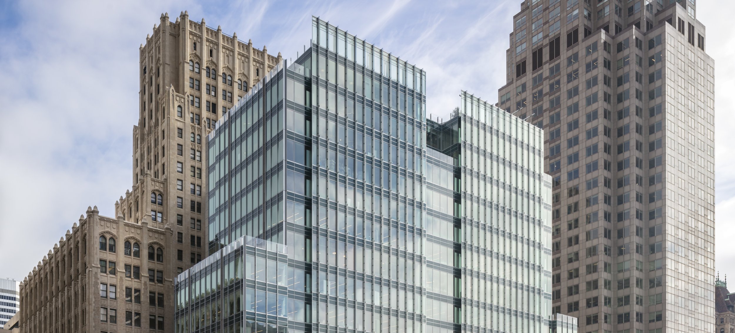 Cityscape with modern glass building flanked by older stone and concrete skyscrapers under cloudy sky.