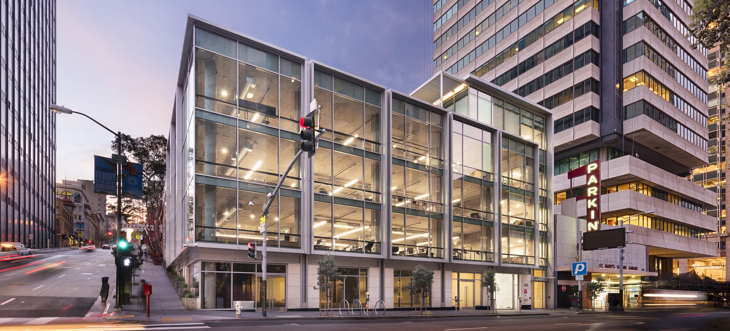 Modern glass office building on a city street during dusk, with a parking lot sign, traffic lights, and light trails from passing vehicles.