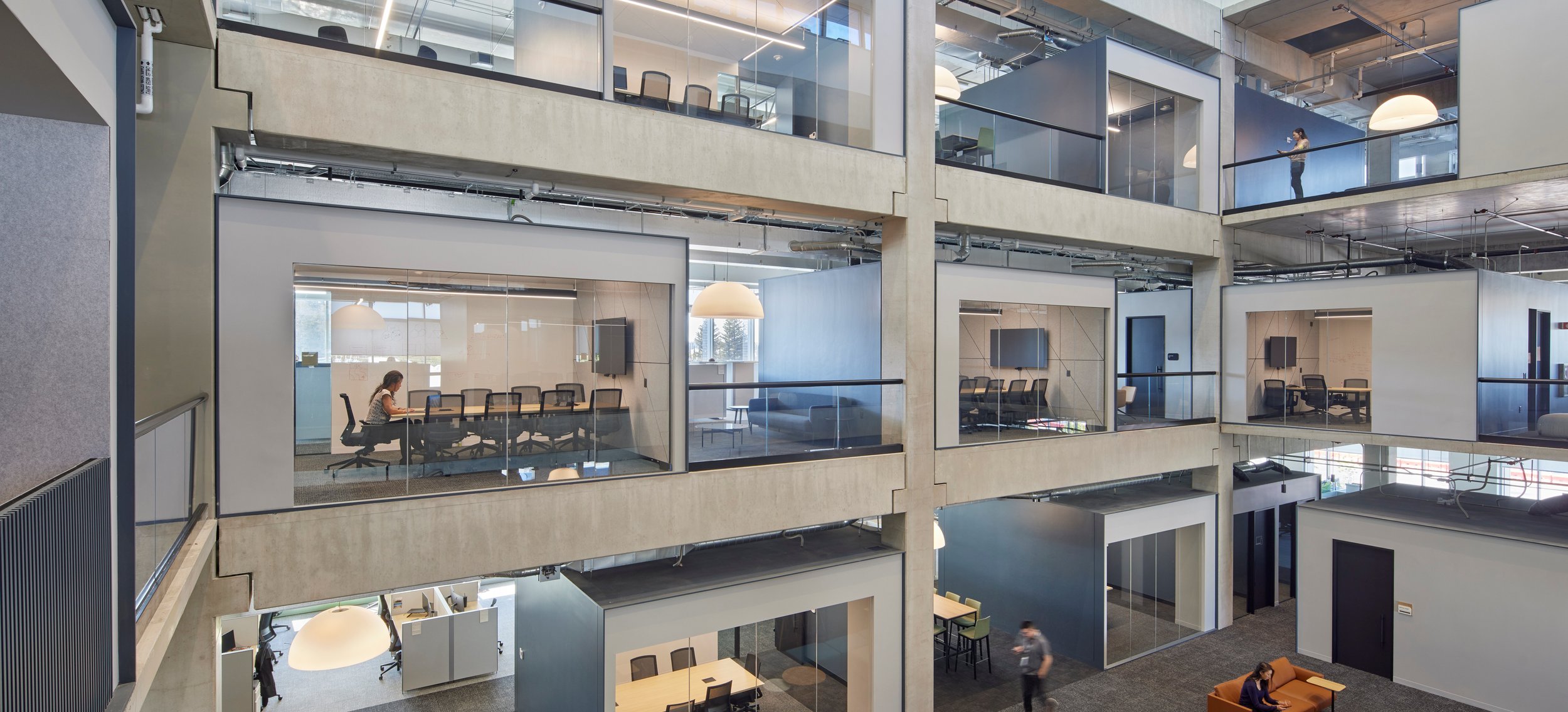 Interior of a modern multi-story office building with glass-walled conference rooms, people working at desks, and a woman standing on an upper balcony looking at her phone.