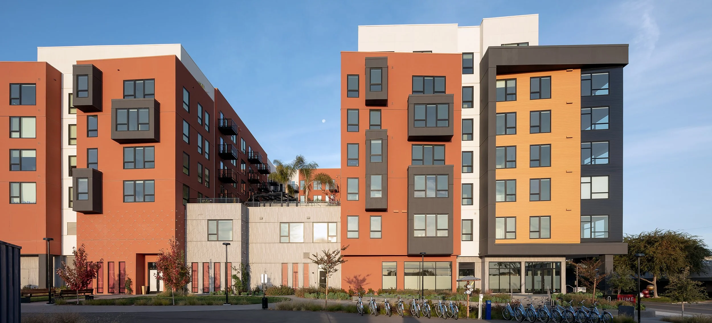 Modern multi-story apartment building with orange, white, and black facades, large windows, and small balconies, set against a blue sky with a full moon, with bike racks and landscaped greenery in the foreground.