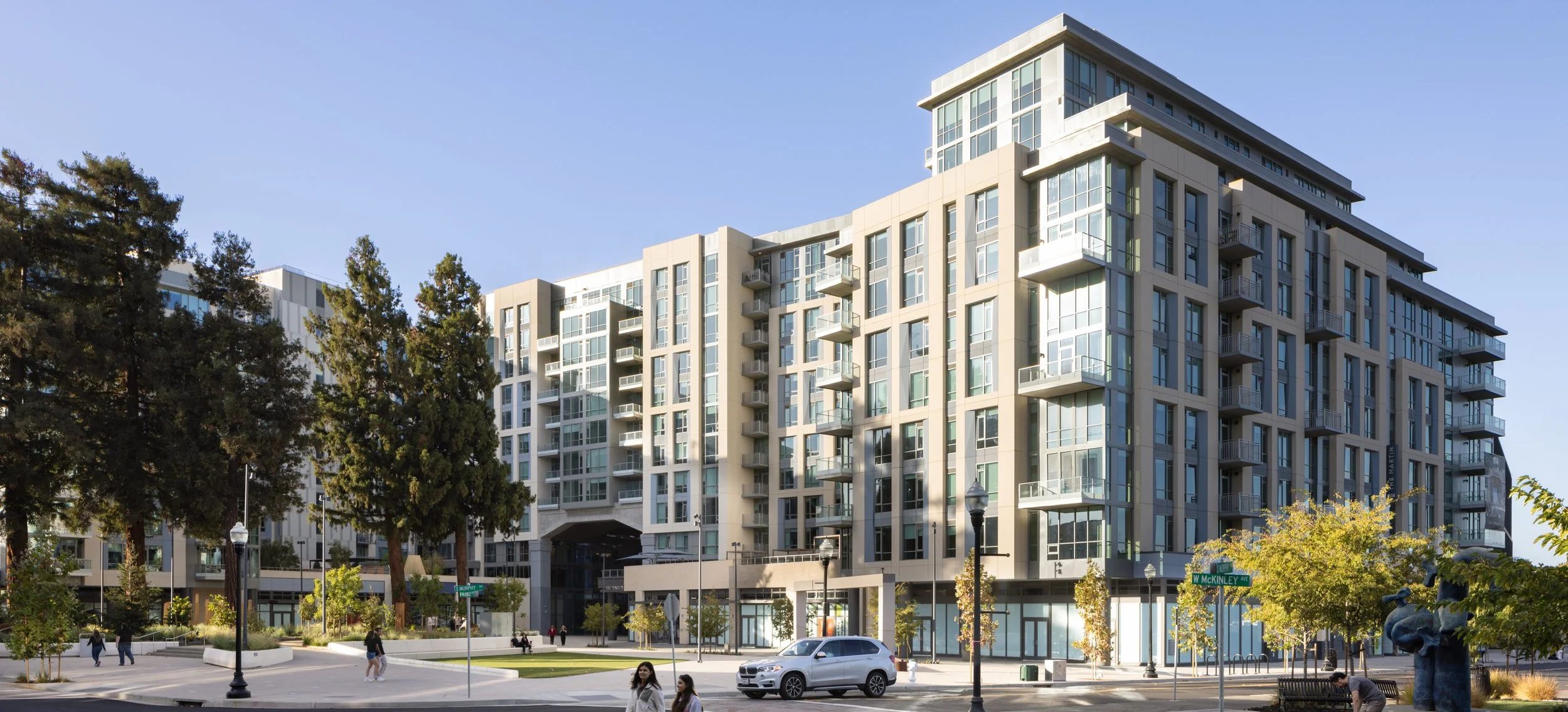 Modern multi-story apartment building with balconies, large glass windows, surrounded by trees and a small park, with pedestrians and parked cars in front.