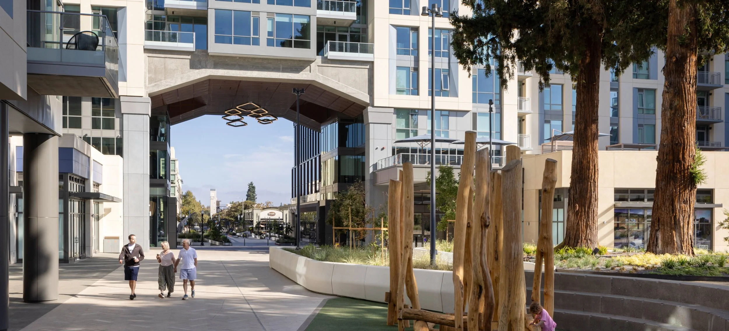 Modern urban courtyard with tall glass buildings, a walkway with people walking, trees, and wooden decorative posts.