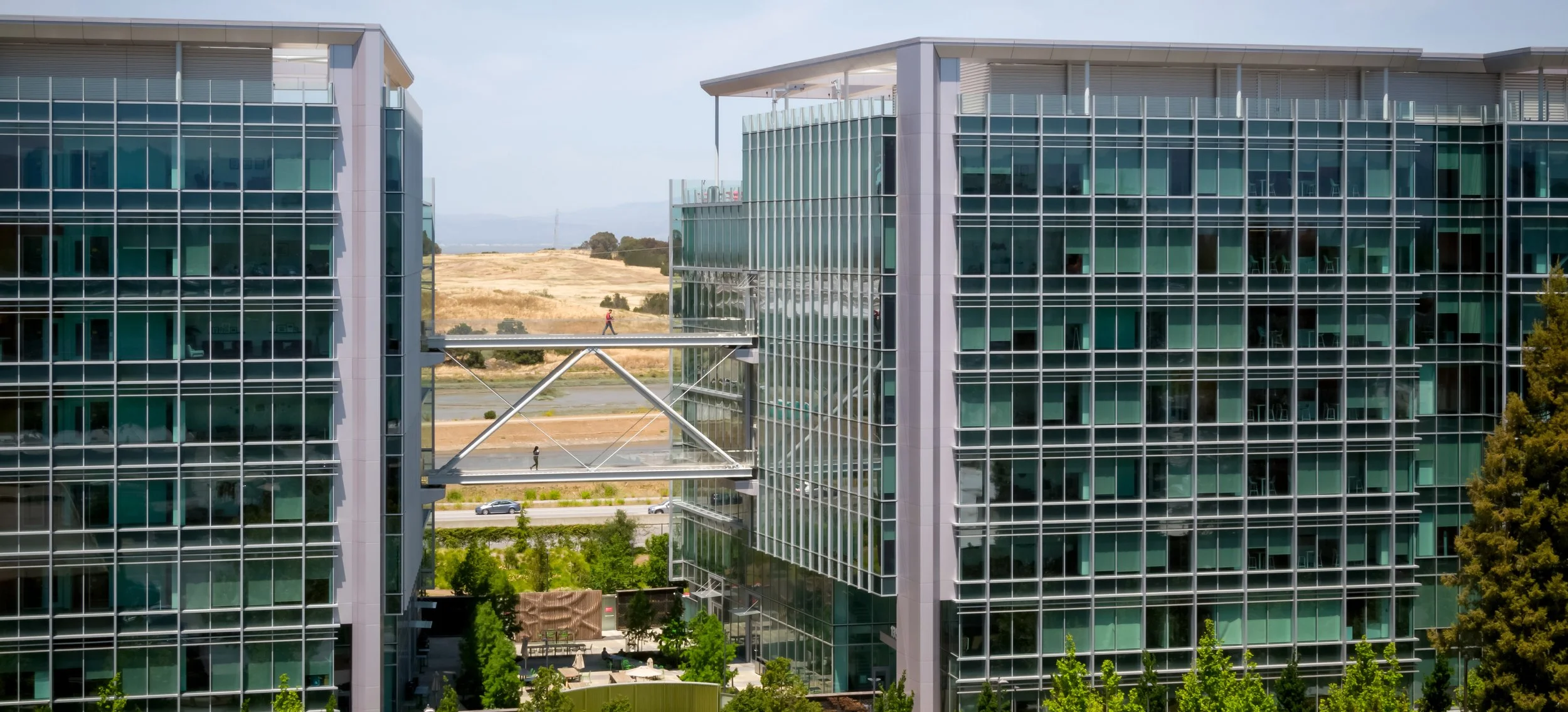 Modern glass office buildings connected by an elevated bridge with pedestrians, set against a backdrop of open landscape and blue sky.
