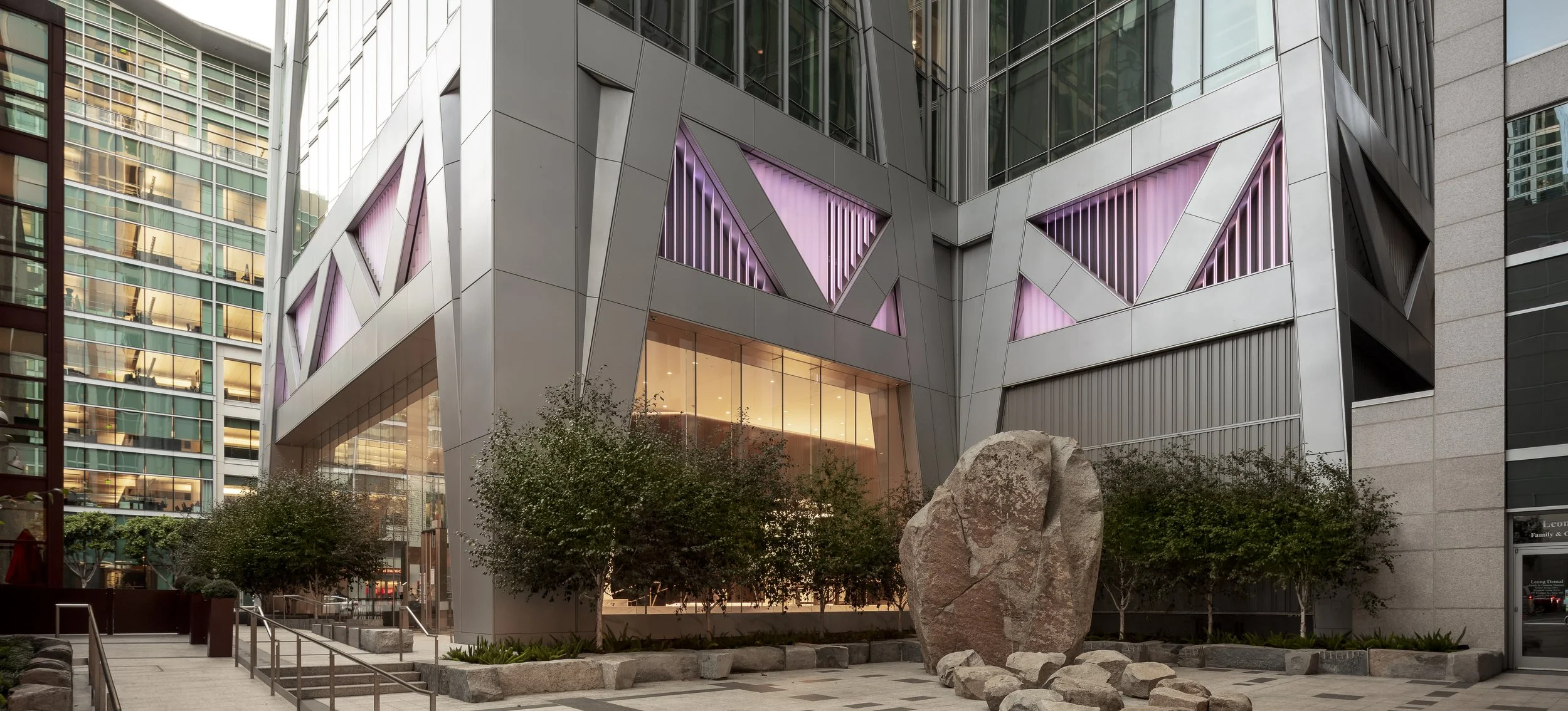 Modern office building with angular design, pink accent lighting in triangular window panels, large glass windows, small landscaped trees, and a rock centerpiece in the foreground.