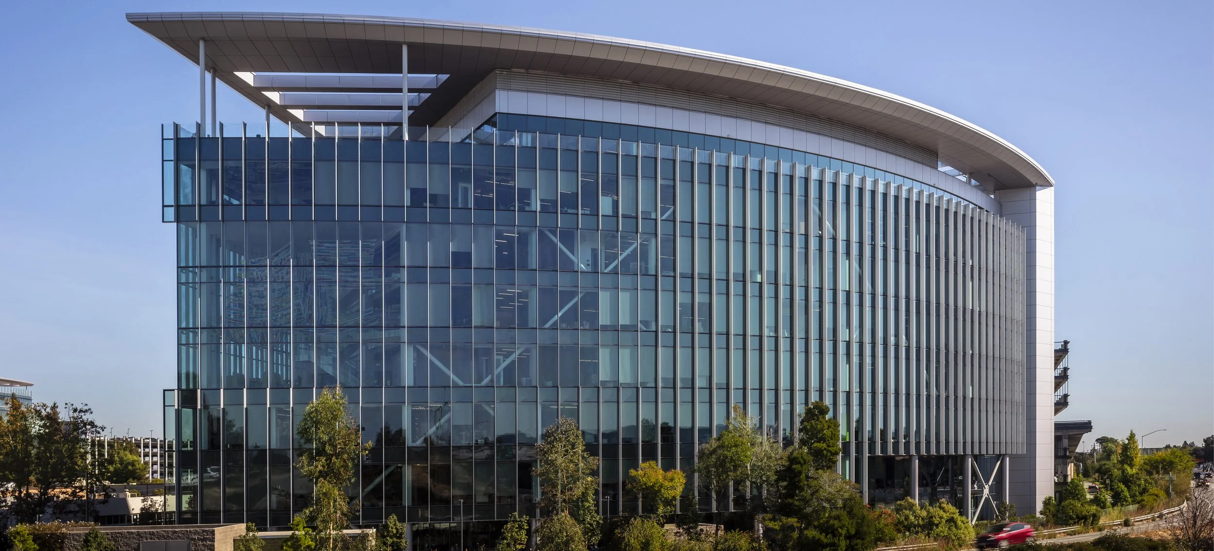 Modern office building with glass facade, vertical metal fins, and a large clock visible through the glass, surrounded by trees and a clear blue sky.
