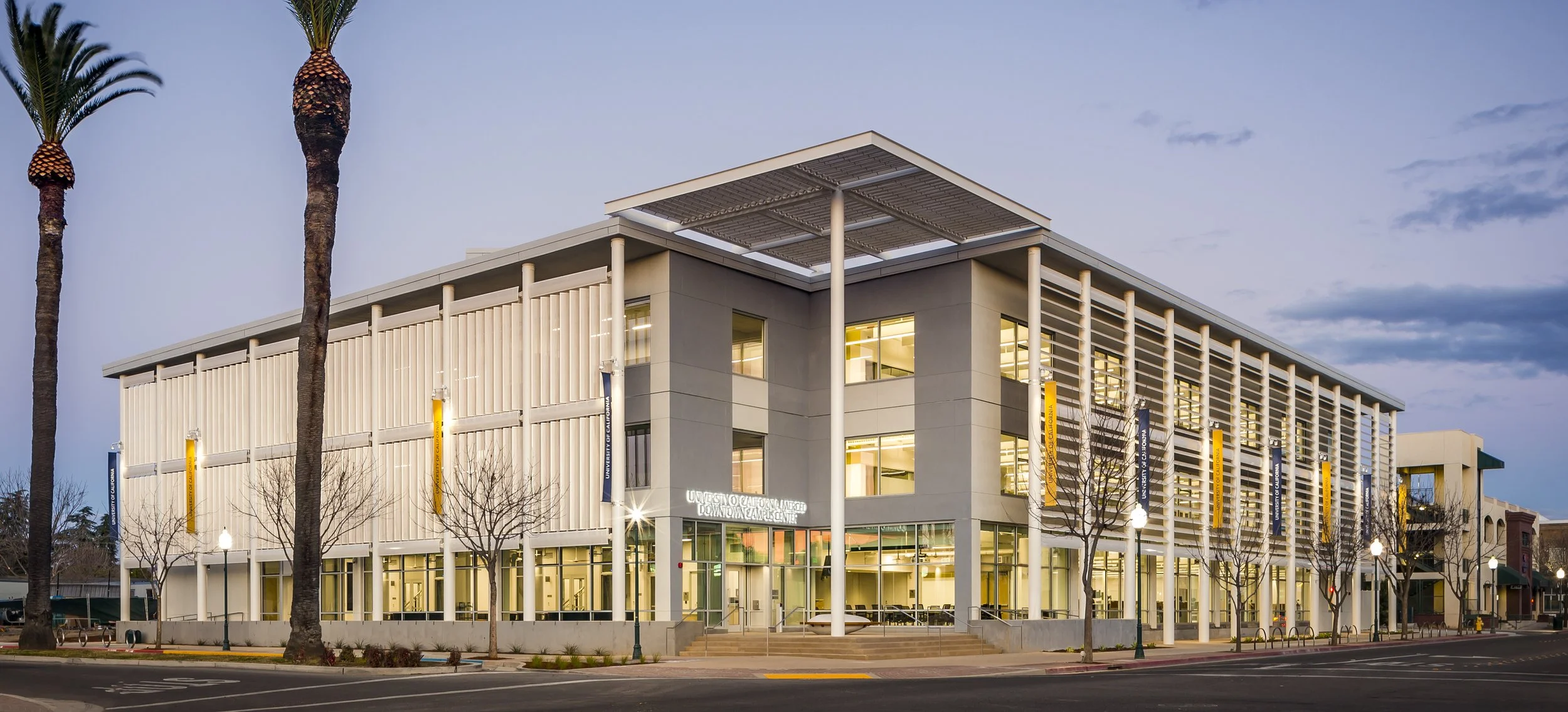 Exterior of a modern university building at dusk with tall palm trees in front, lit windows, and banners on the façade reading 'University of California, Merced, Downtown Campus Center'.