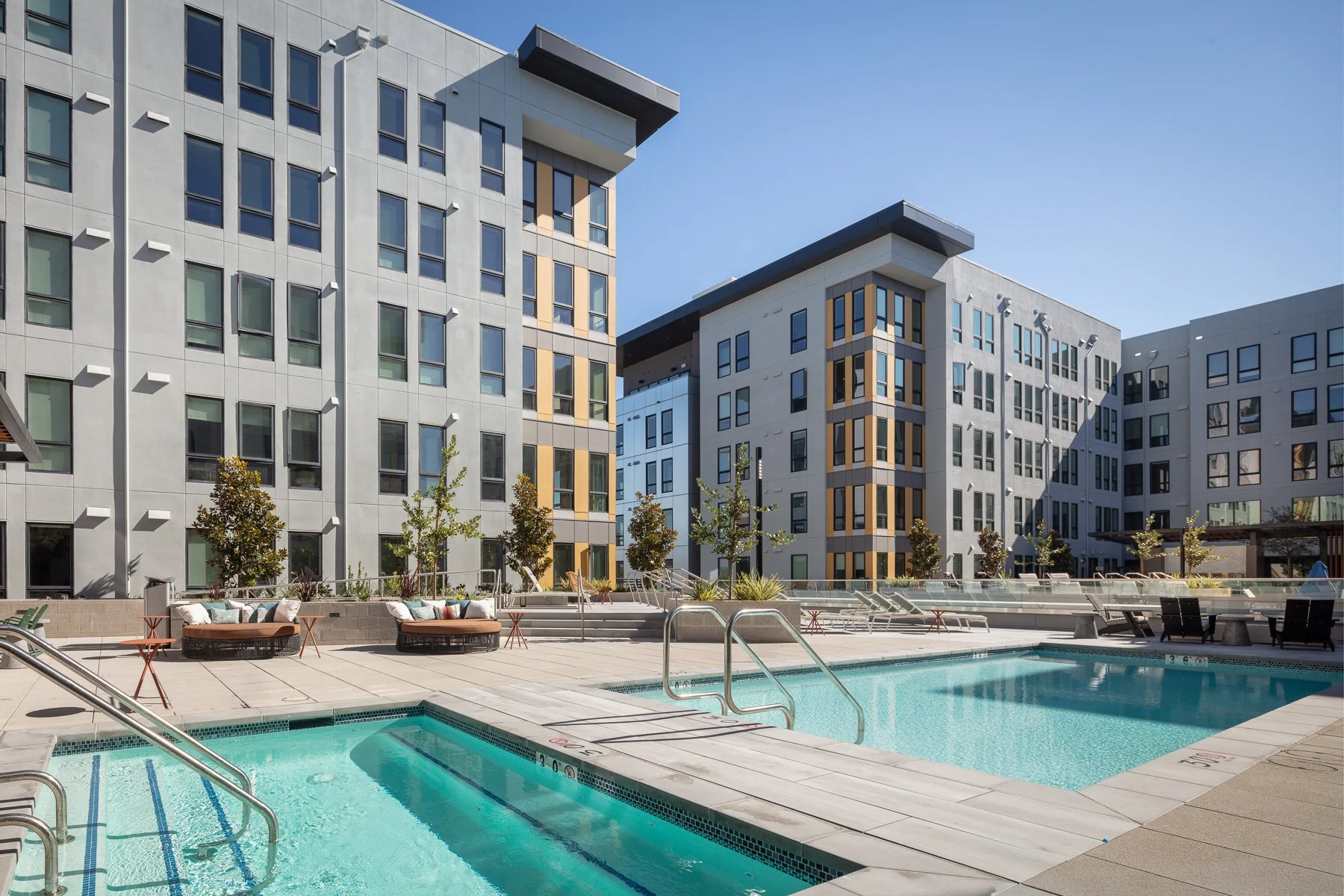 Modern apartment complex with an outdoor pool and lounge chairs, surrounded by residential buildings, under a clear blue sky.