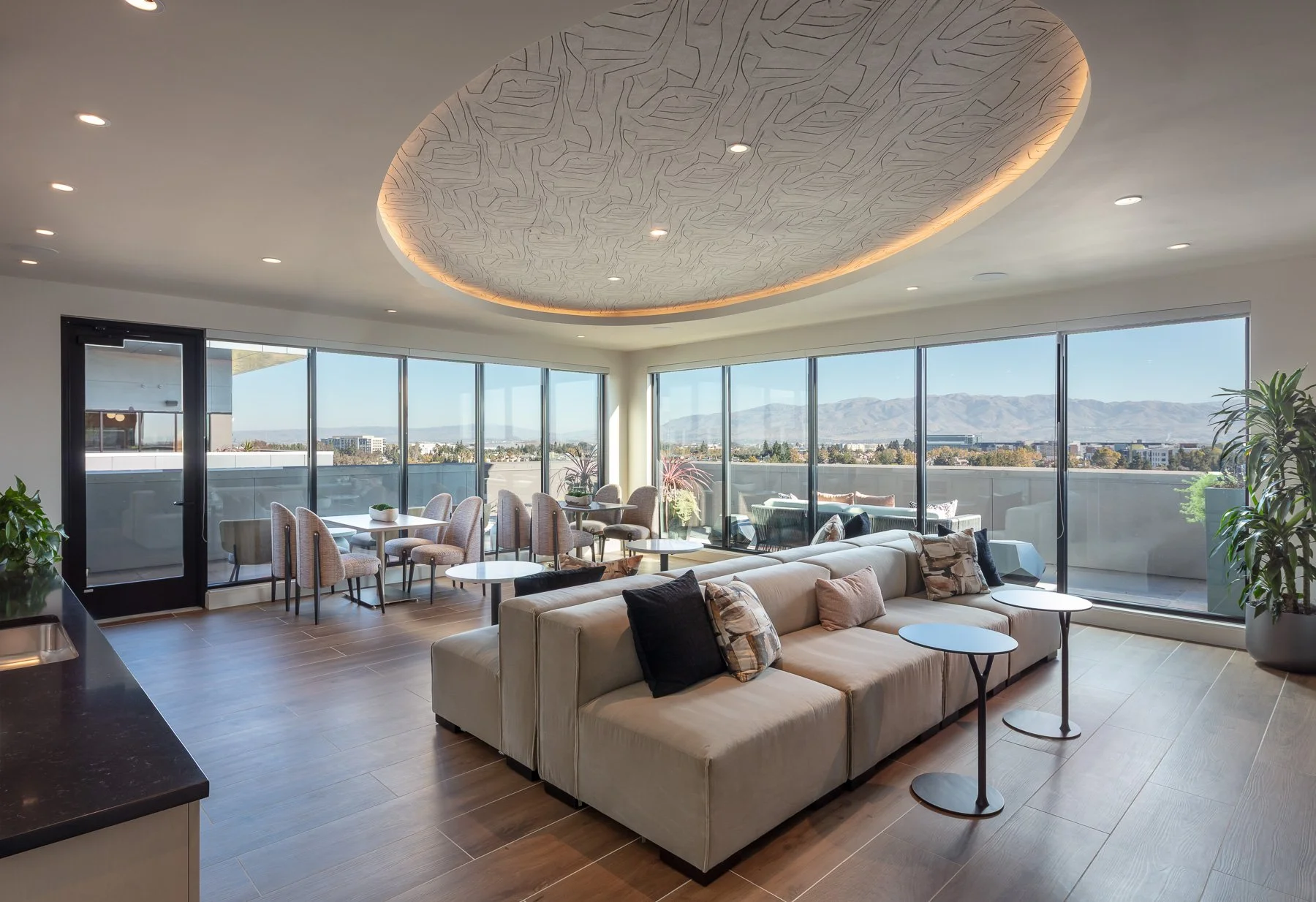 Modern living room with large floor-to-ceiling windows overlooking city and mountains, featuring a white sectional sofa with pillows, small side tables, and a dining nook with chairs and tables near the windows.