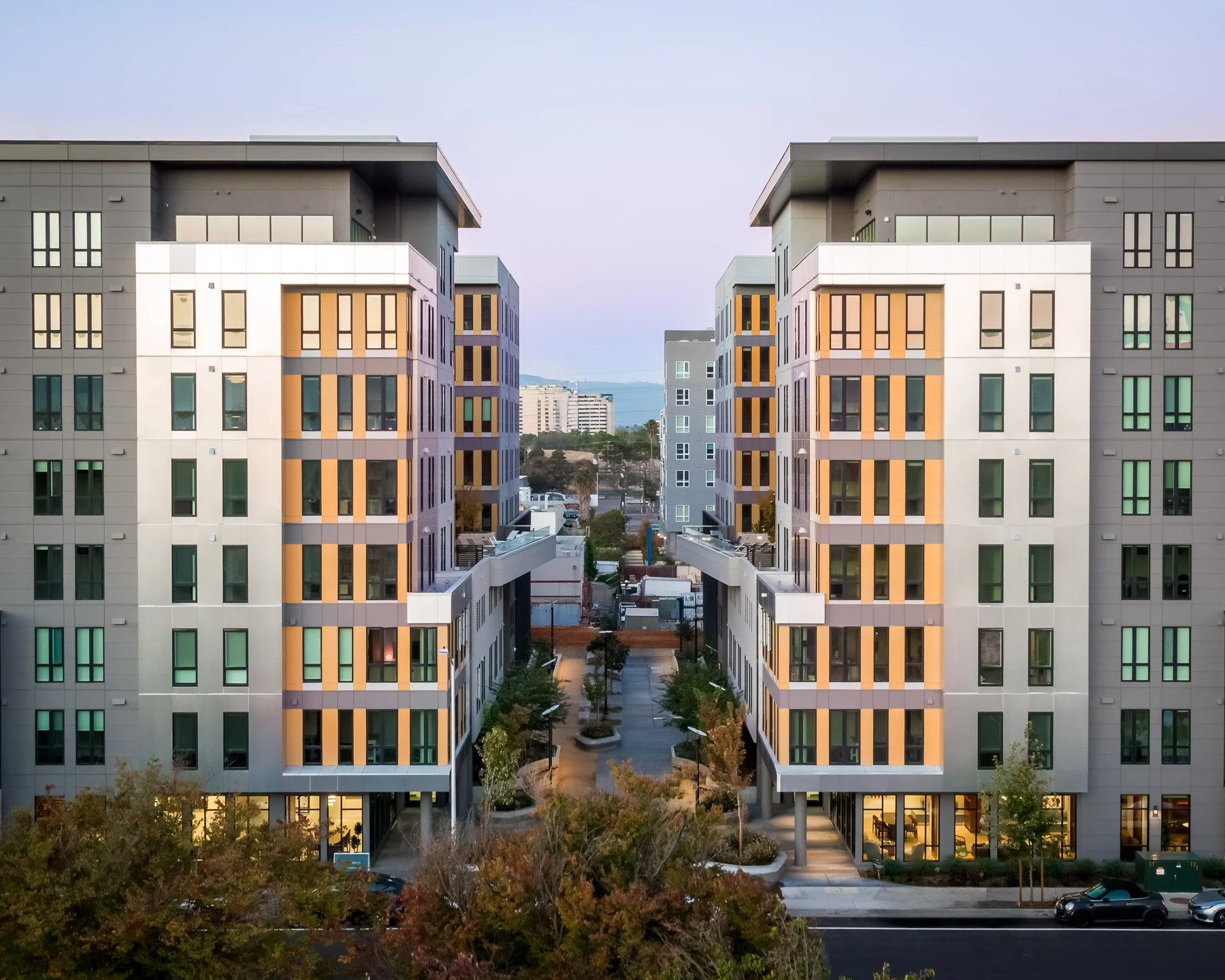 A modern residential apartment complex with two symmetrical buildings separated by a courtyard, featuring large windows and colorful accents.