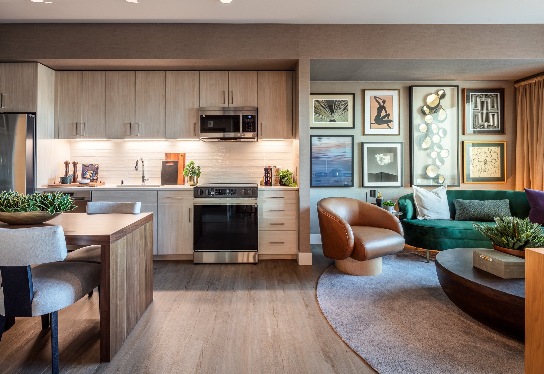 Open-concept living room and kitchen in a modern apartment with light wood finishes, green velvet couch, brown leather chair, and assorted wall art.