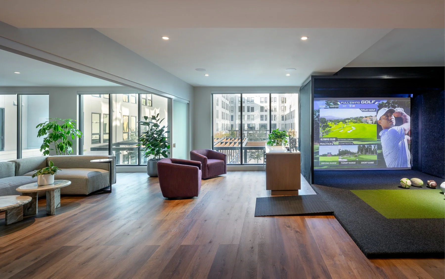 A modern indoor lounge area with large windows, green plants, beige and maroon chairs, and a large television displaying a golf game.