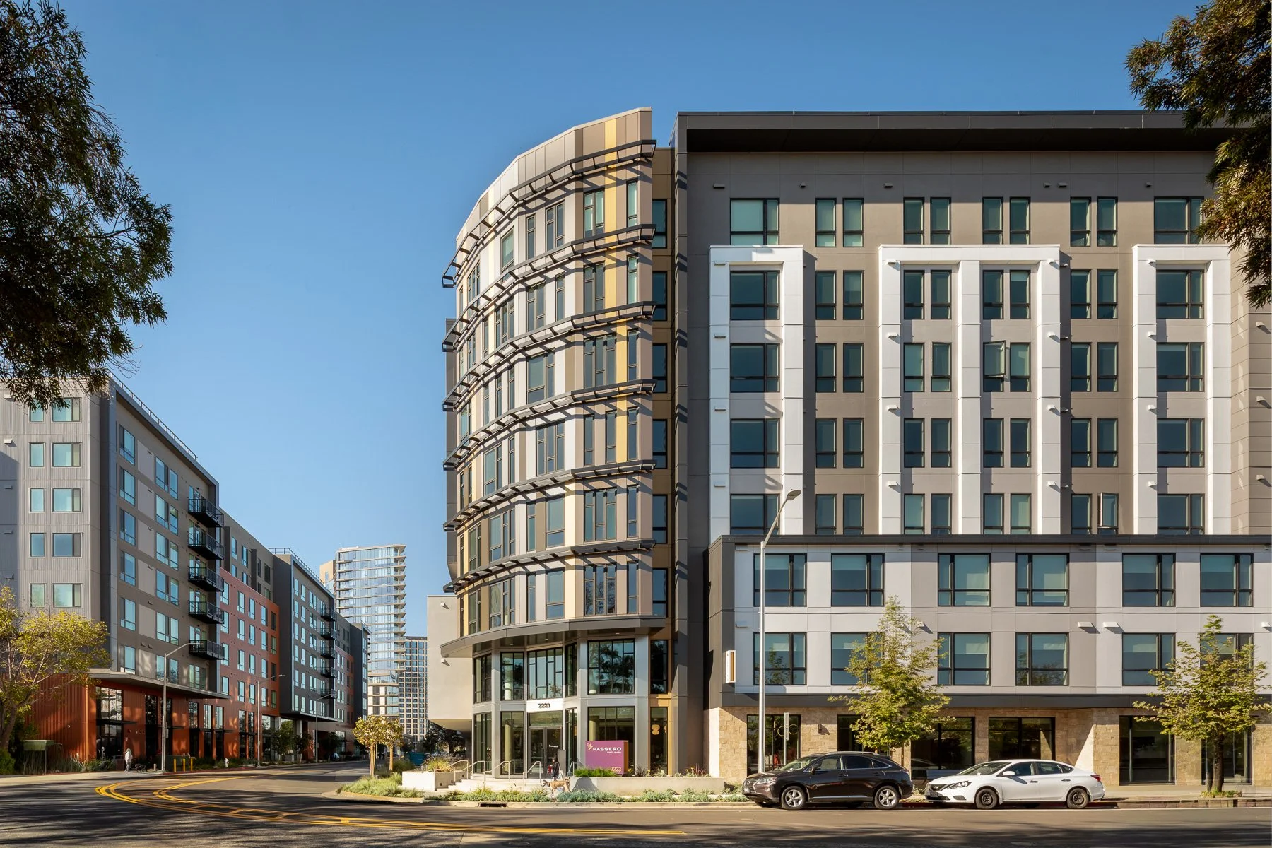Modern multi-story residential or hotel building with large glass windows and a rounded corner. Cars are parked on the street in front, and there are trees and a clear blue sky.