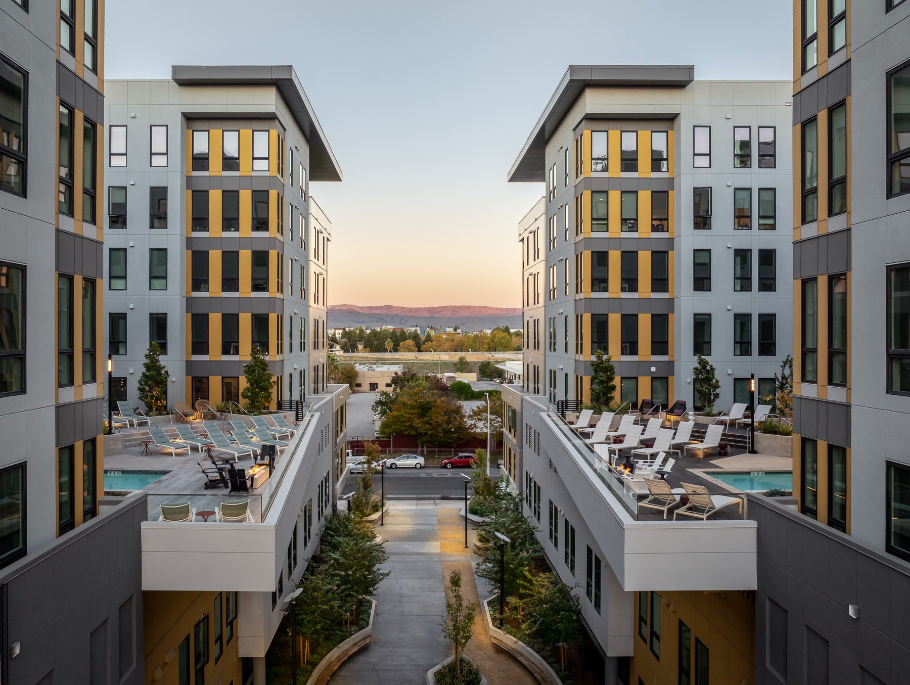 Modern apartment complex with outdoor pools, lounge chairs, and trees at sunset.