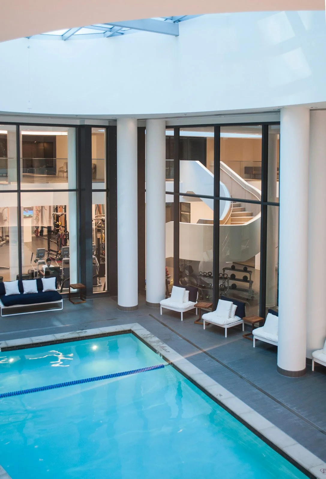 Indoor swimming pool with lounge chairs and large windows overlooking gym area.