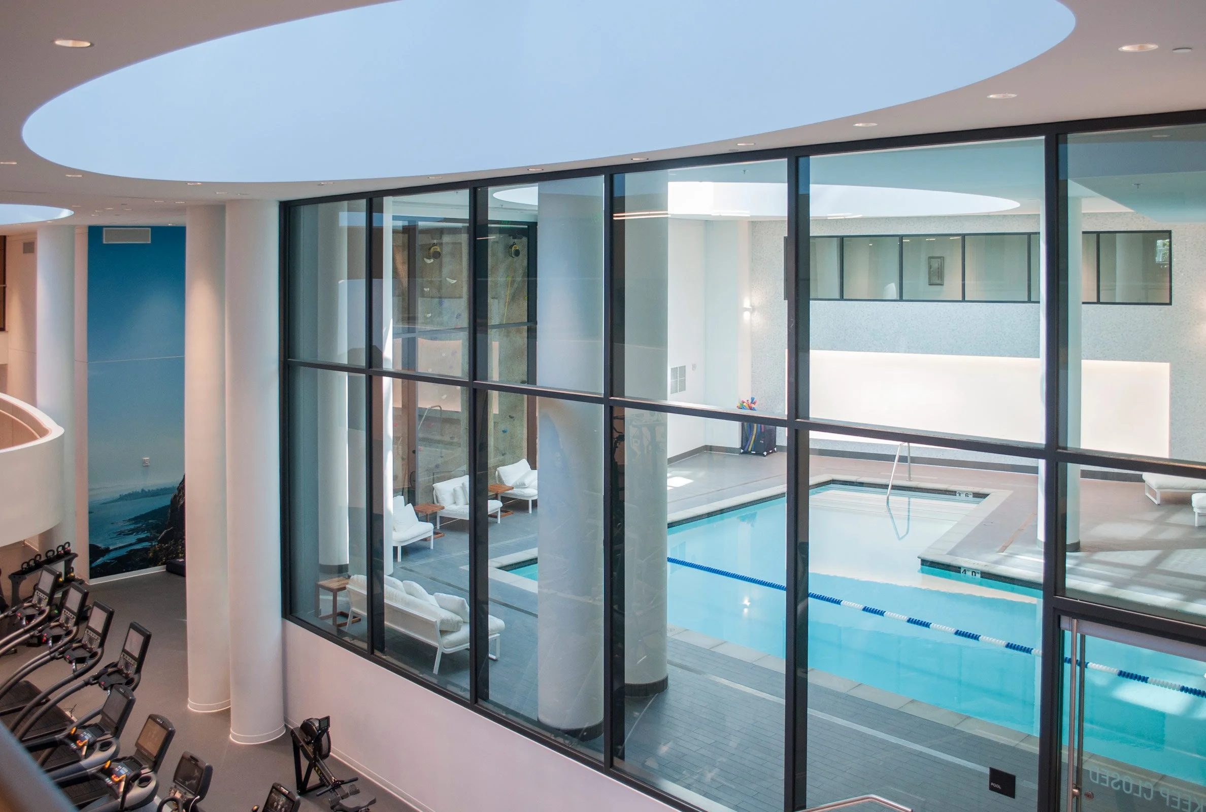 Interior view of a modern indoor swimming pool area seen through large glass windows, with lounge chairs and a small hot tub or spa beside the pool.