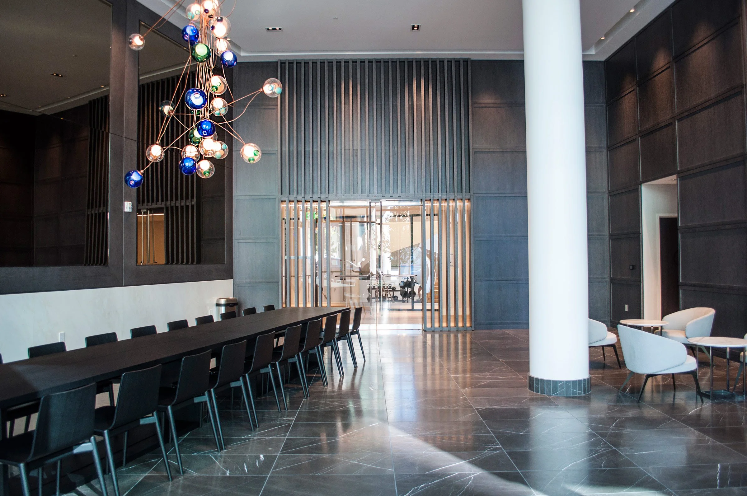 Modern hotel lobby with black wall paneling, white column, black long table with chairs, armchairs with side tables, and colorful pendant light fixture.