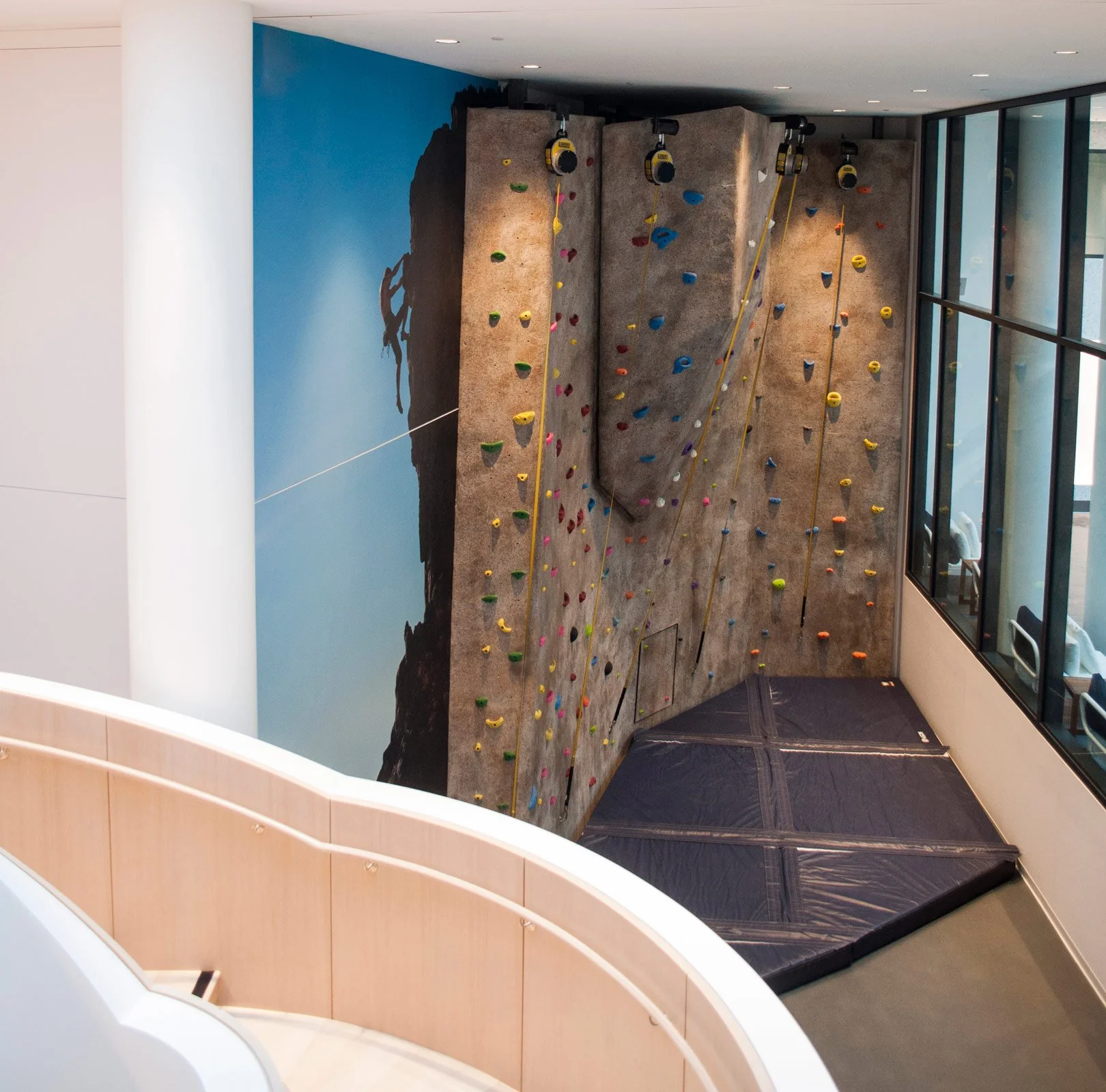 Indoor rock climbing wall with various colored holds and safety mats beneath, next to a large window.