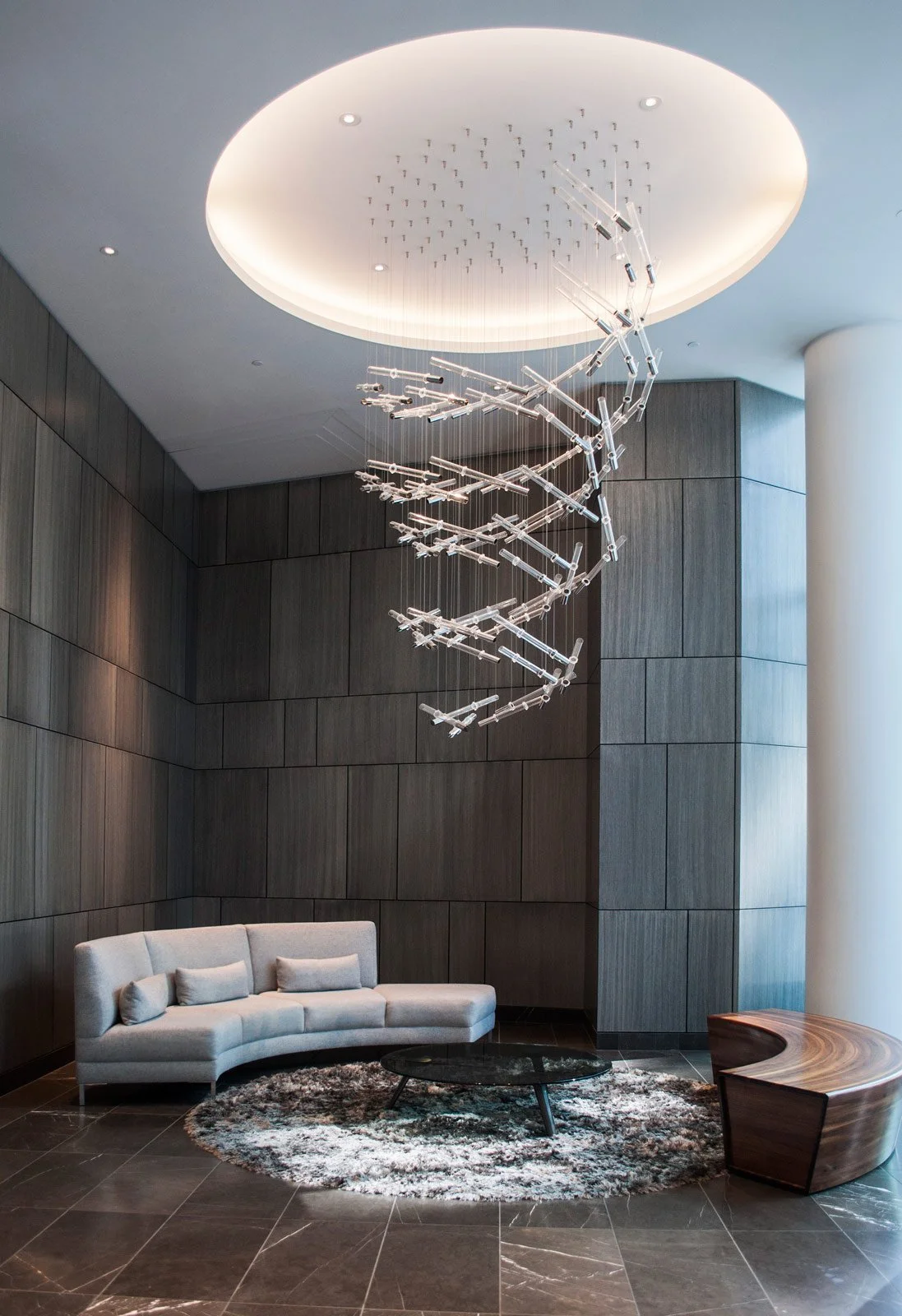 Modern lobby with a white curved sofa, a round table, and a wooden bench beneath a large spiral chandelier in a high-ceiling room with dark wood-paneled walls and tile flooring.