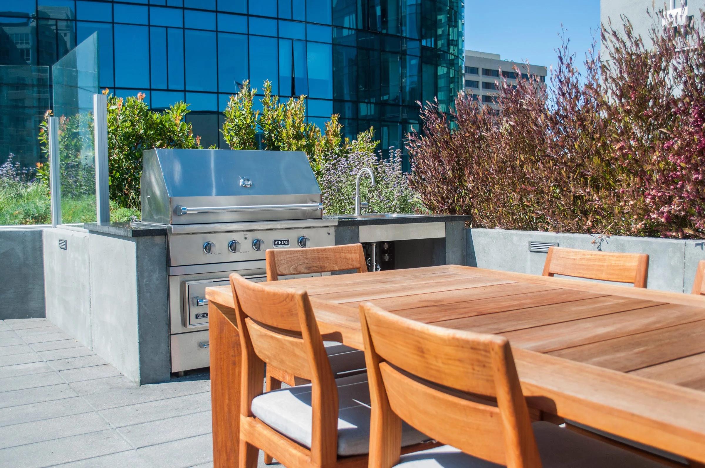 Outdoor patio area with wooden dining table and chairs, stainless steel grill, sink, and lush greenery in an urban setting with high-rise buildings and a blue sky.