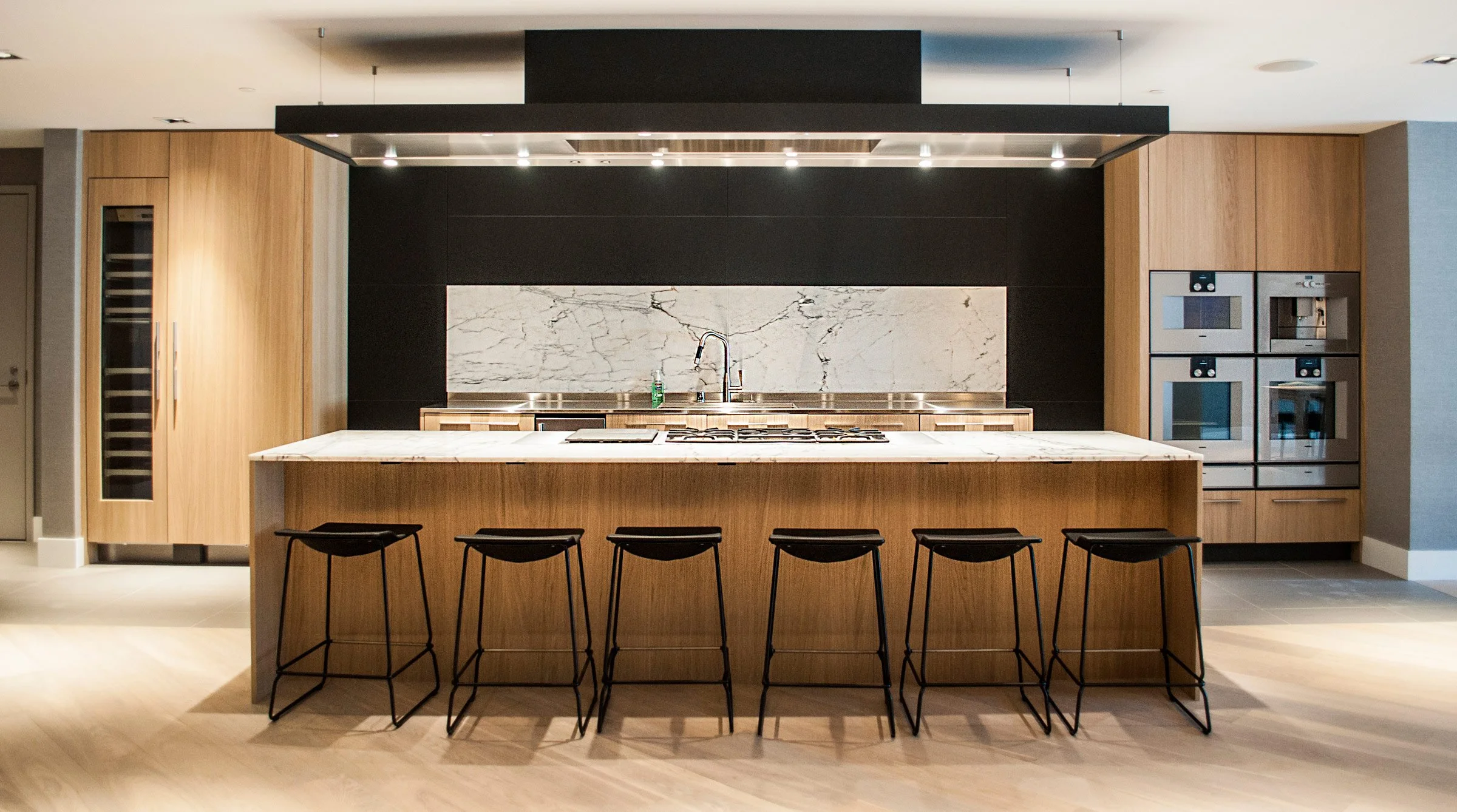 Modern kitchen with a central island with black bar stools, a marble countertop, wooden cabinets, stainless steel appliances, and a black and white marble backsplash.
