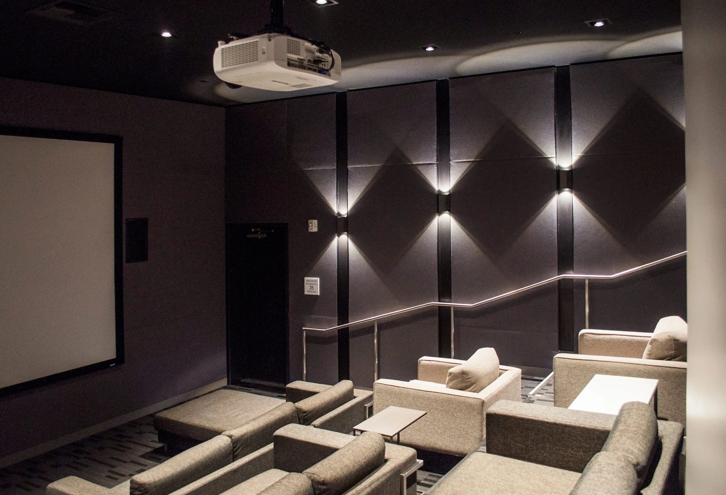 Empty movie theater with multiple beige armchairs facing a large blank screen, dark purple walls with geometric light fixtures, and a ceiling-mounted projector.