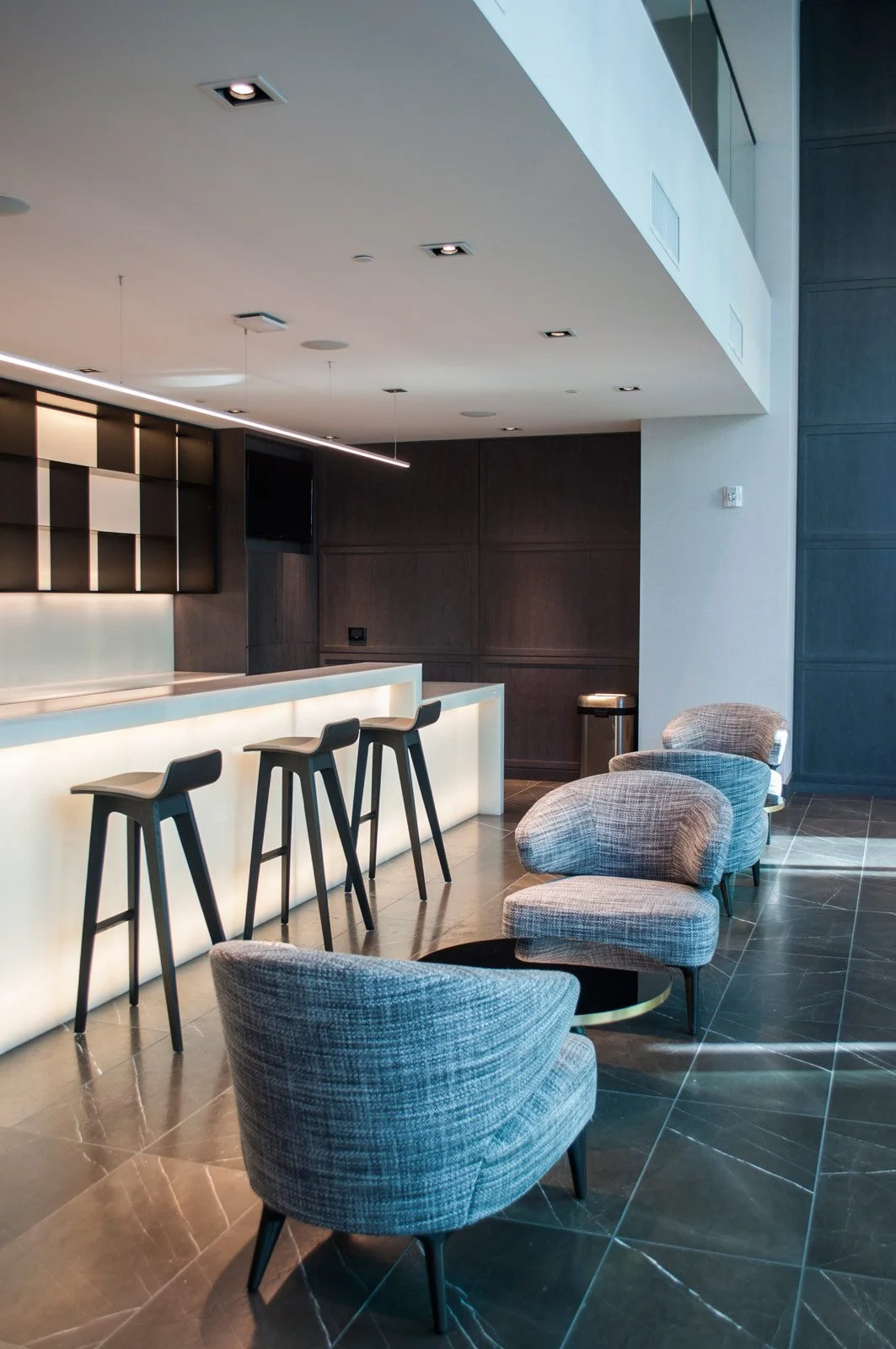 Modern hotel lobby with a bar area, high stools, and plush armchairs, illuminated by recessed ceiling lights and natural light.
