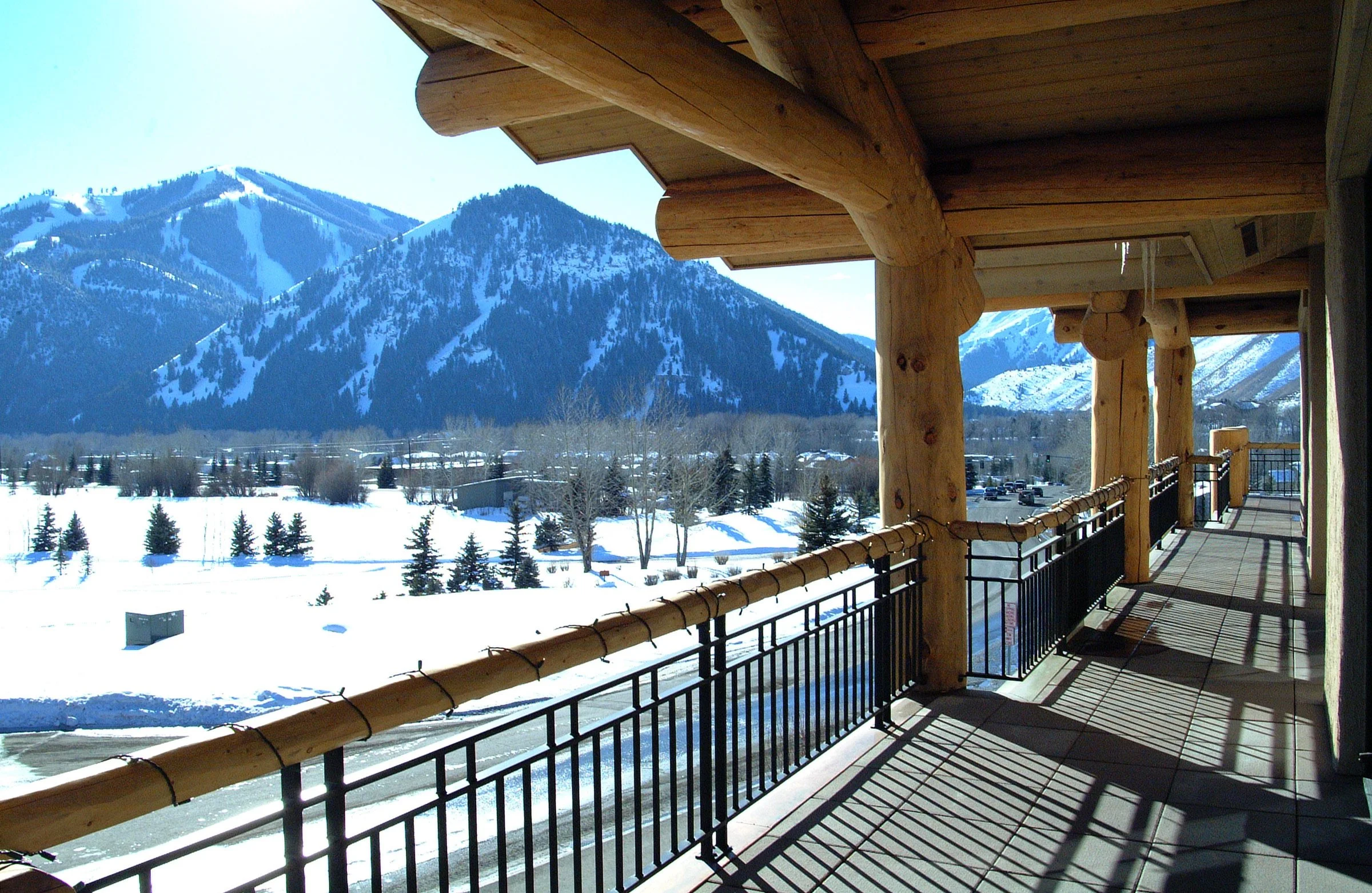 Snow-covered landscape with mountains, trees, and a balcony with wooden beams and black iron railing.