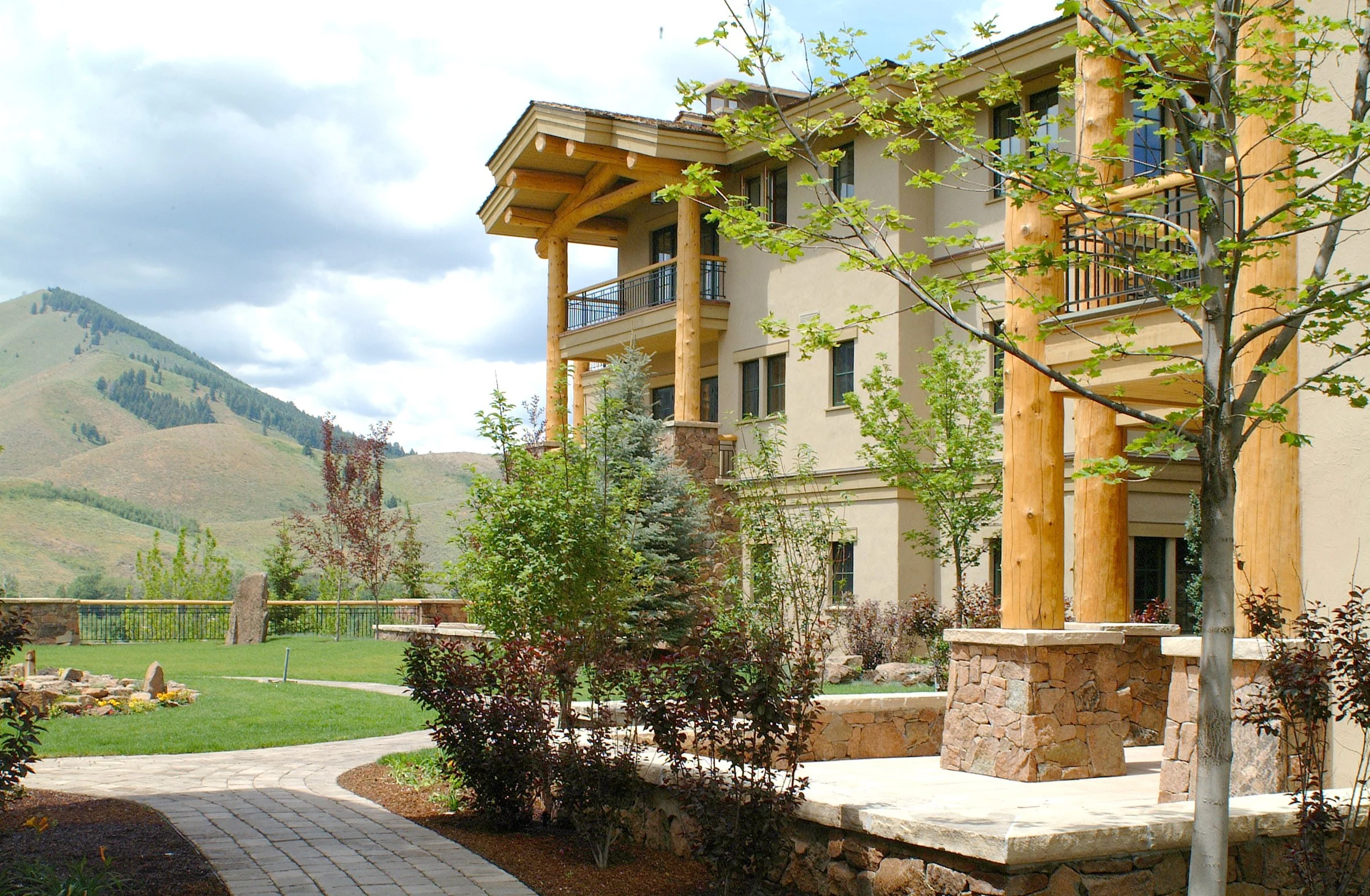 Exterior view of a modern multi-story residential building with wooden columns and balconies, surrounded by a landscaped yard with trees, shrubs, a brick pathway, and mountains in the background.