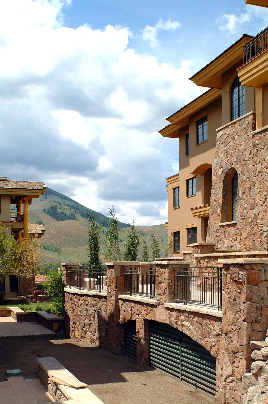 View of multi-story residential buildings with stone and stucco exteriors, set against a backdrop of green hills and partly cloudy sky.