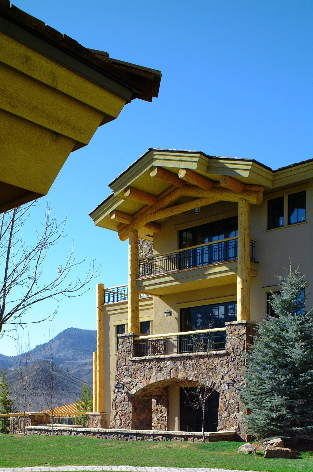 A multi-story house with a stone and stucco exterior and wooden beams on the balcony, situated in a mountainous area with trees and a grassy lawn.