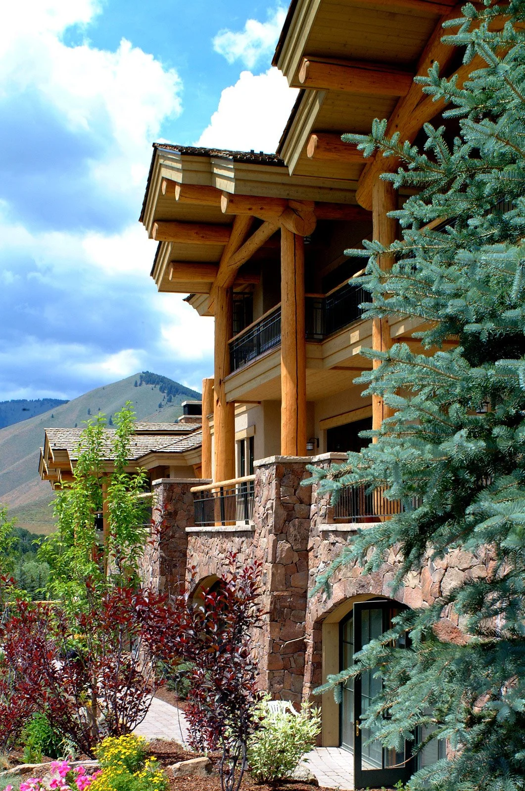 A multi-story mountain lodge with wooden beams, stone accents, and balconies, surrounded by greenery and set against mountain terrain with a partly cloudy sky.