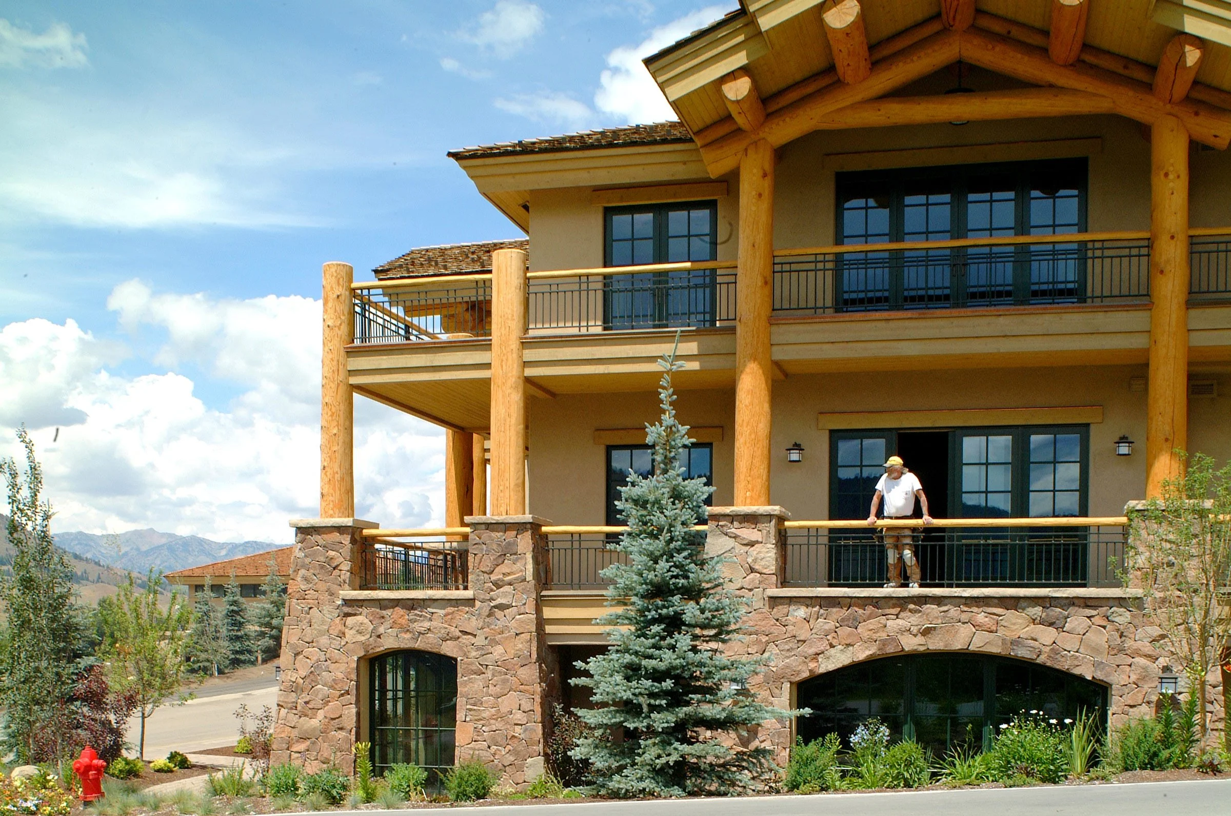 A two-story house with stone and wood exterior, surrounded by landscaping, with a man standing on the front porch railing, and mountains in the background under a partly cloudy sky.