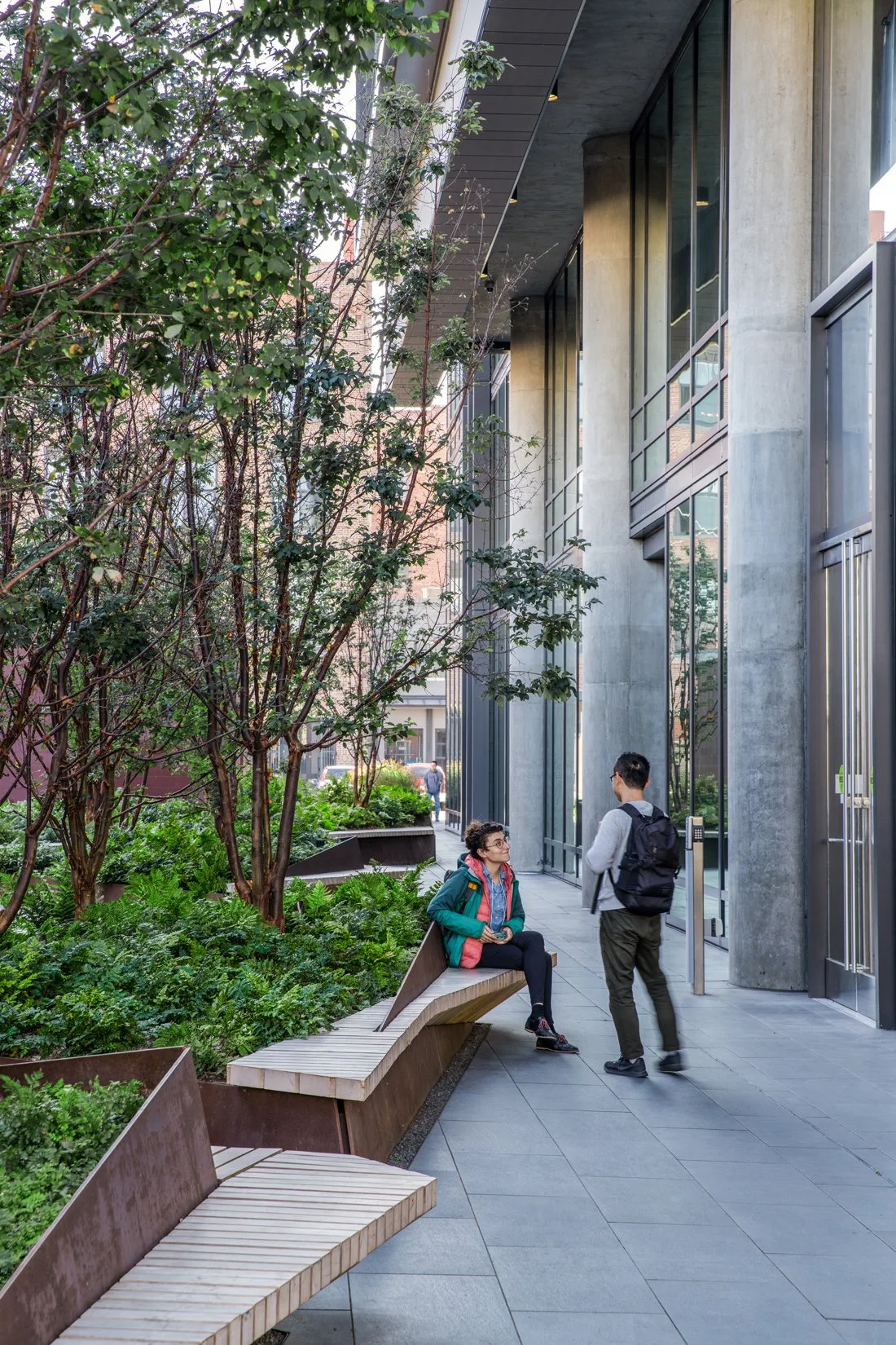 A woman sitting on a curved wooden bench talking to a man standing beside her in front of modern glass building with greenery and trees around.