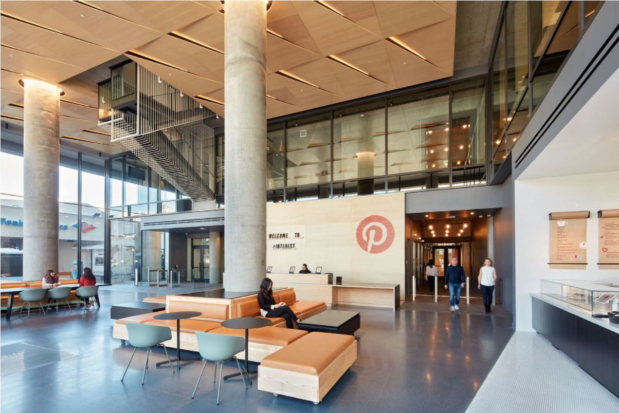 Modern lobby of a Pinterest office building with a reception desk, seating area with orange sofas, a few people sitting and walking, large glass windows, and high ceiling with wooden panels.