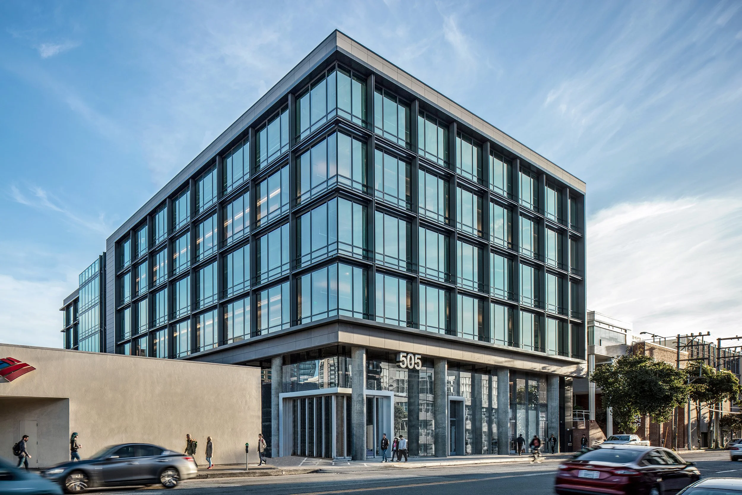 Modern glass office building at 505 on a city street with cars and pedestrians.