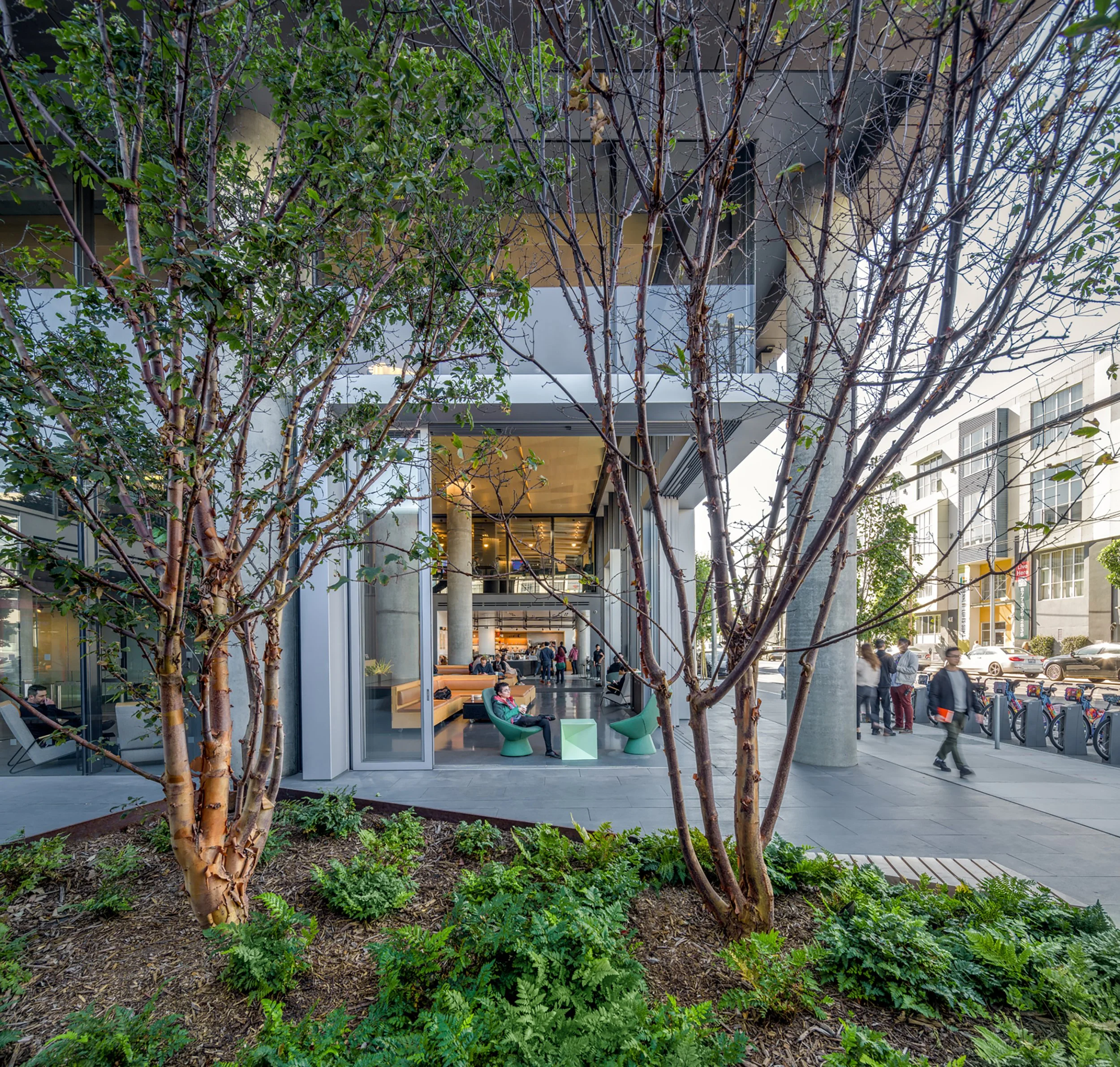 Urban sidewalk view with trees, a modern building entrance, and people walking or sitting outside; indoor lobby visible through glass doors.