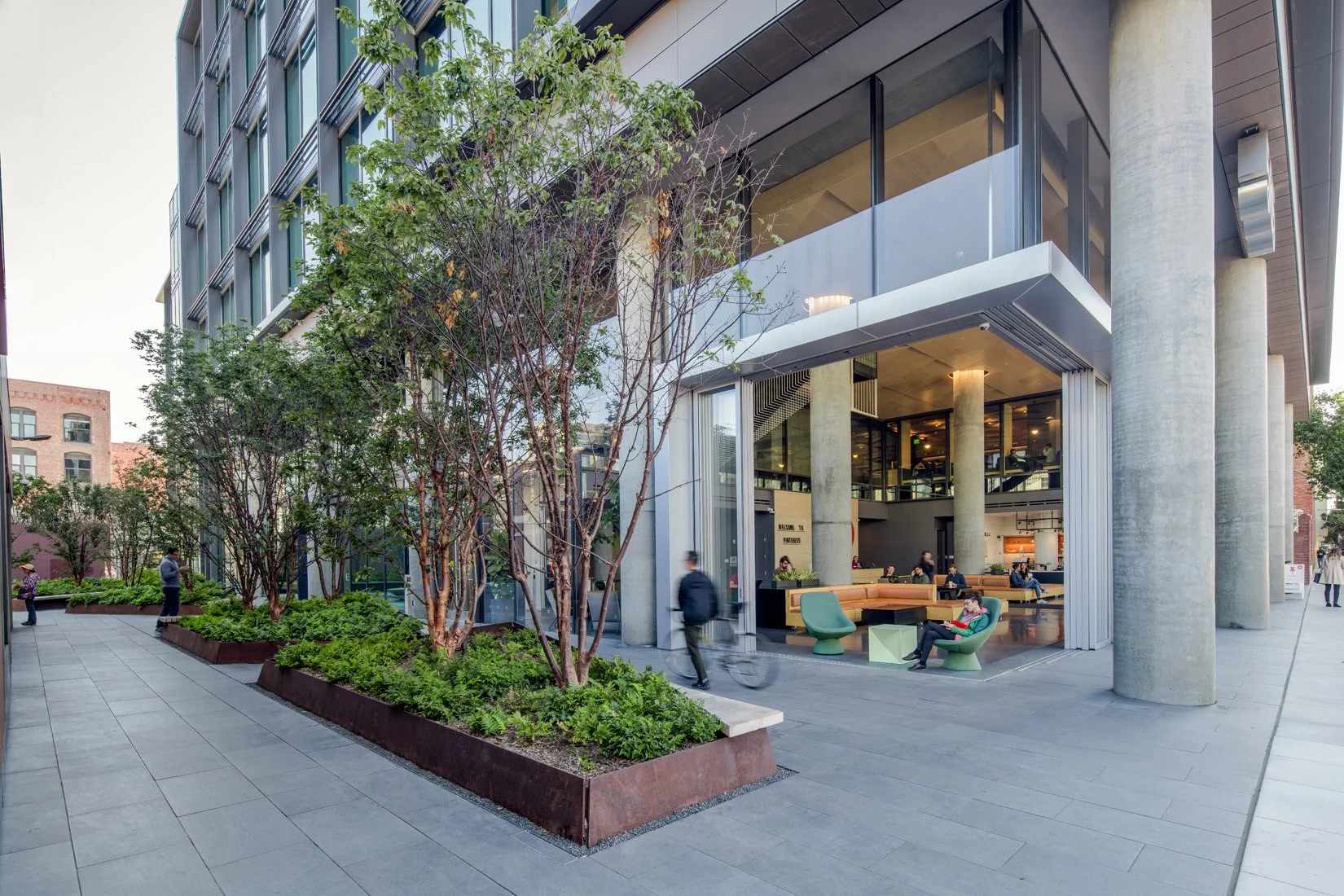 Modern building with large glass windows and concrete columns, outdoor seating area with colorful chairs and tables, green plants and trees in planters, people walking and sitting outside, urban street scene during daytime.