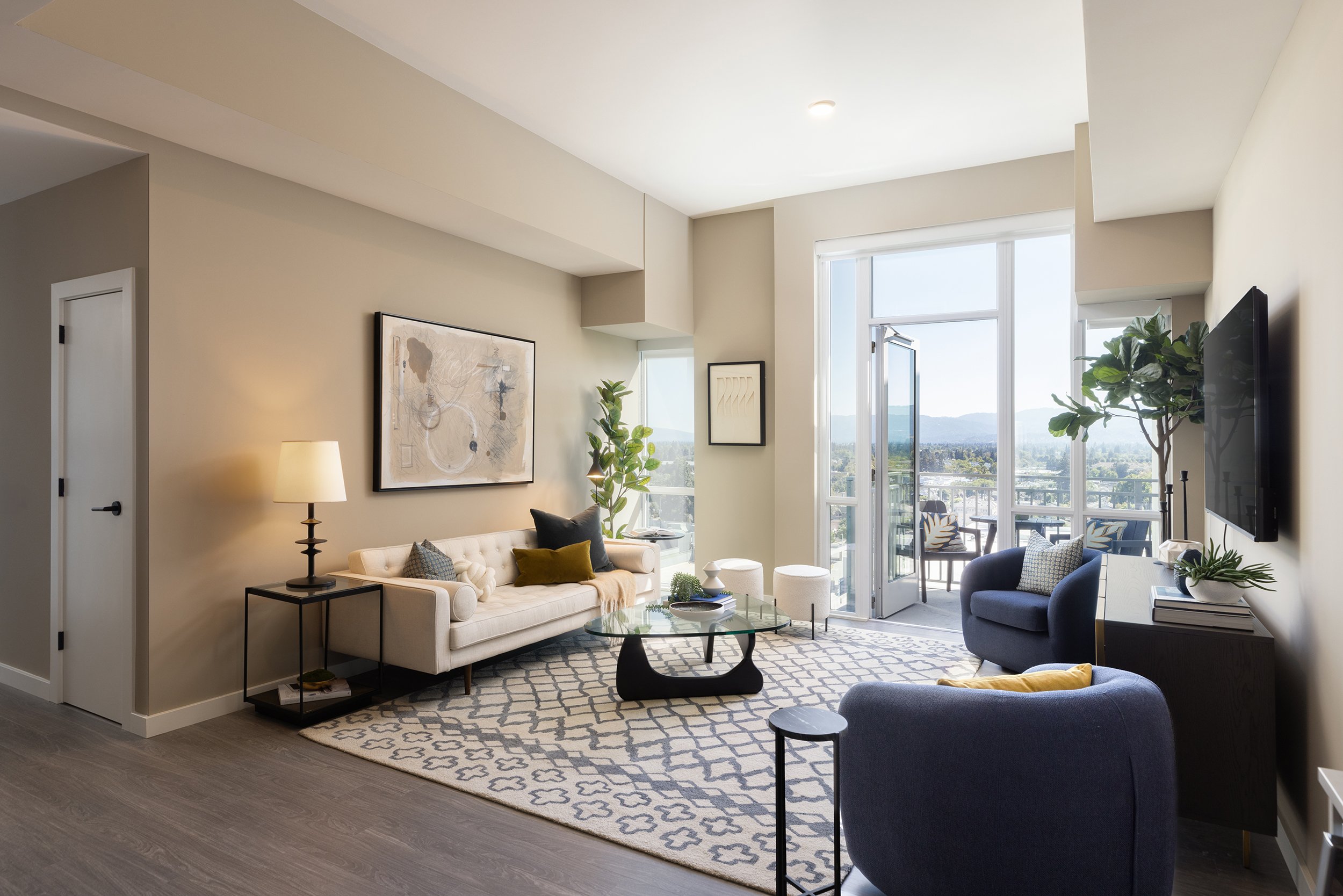Living room with natural light, featuring a white sofa, a glass coffee table, two blue armchairs, a black TV mounted on the wall, and a balcony with outdoor seating.