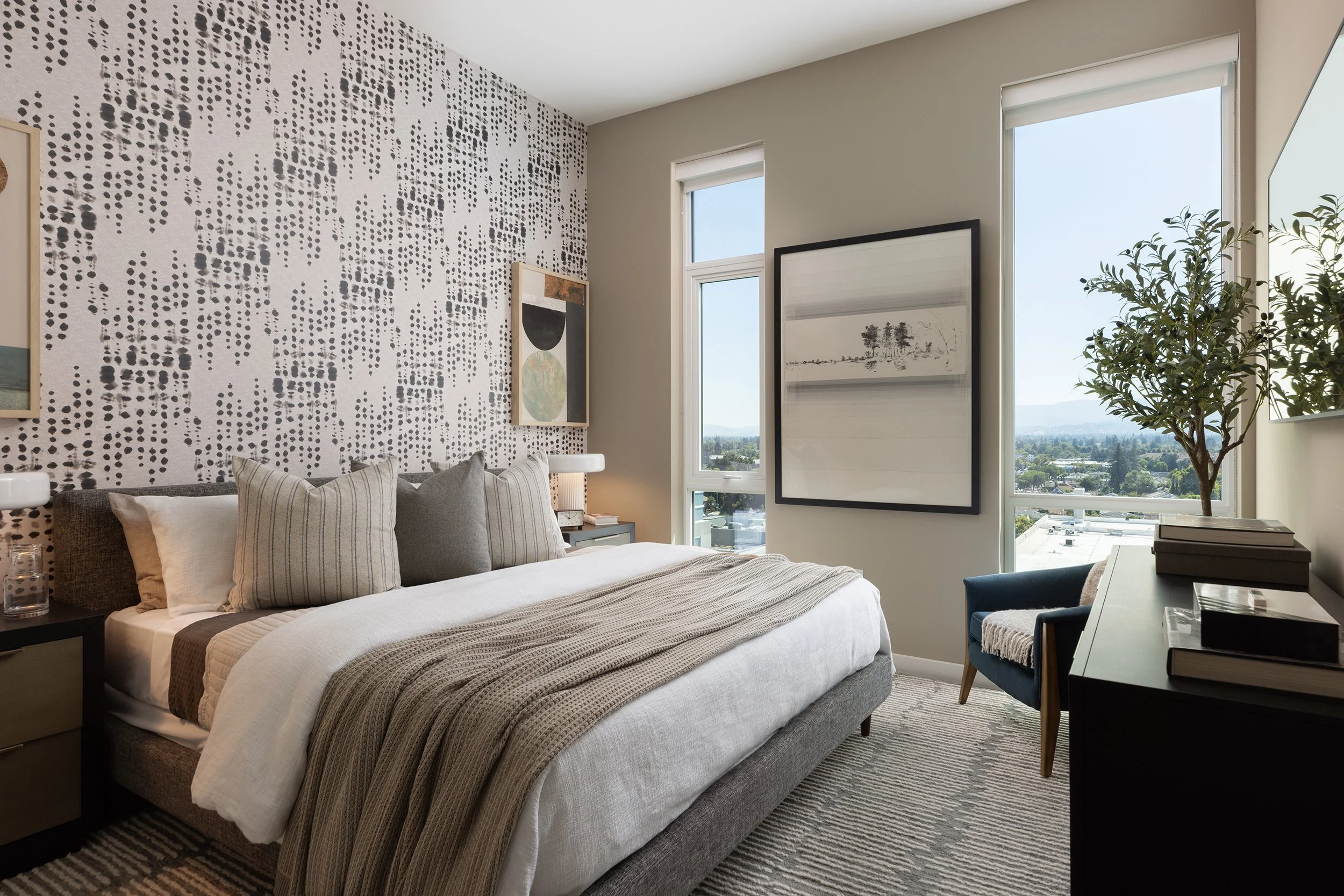 A modern bedroom with a large bed, decorative pillows, and a textured blanket. The room has abstract wall art, a tall window with a city view, a dark dresser with stacked books, a blue armchair, and a large potted plant. The decor is neutral with bla