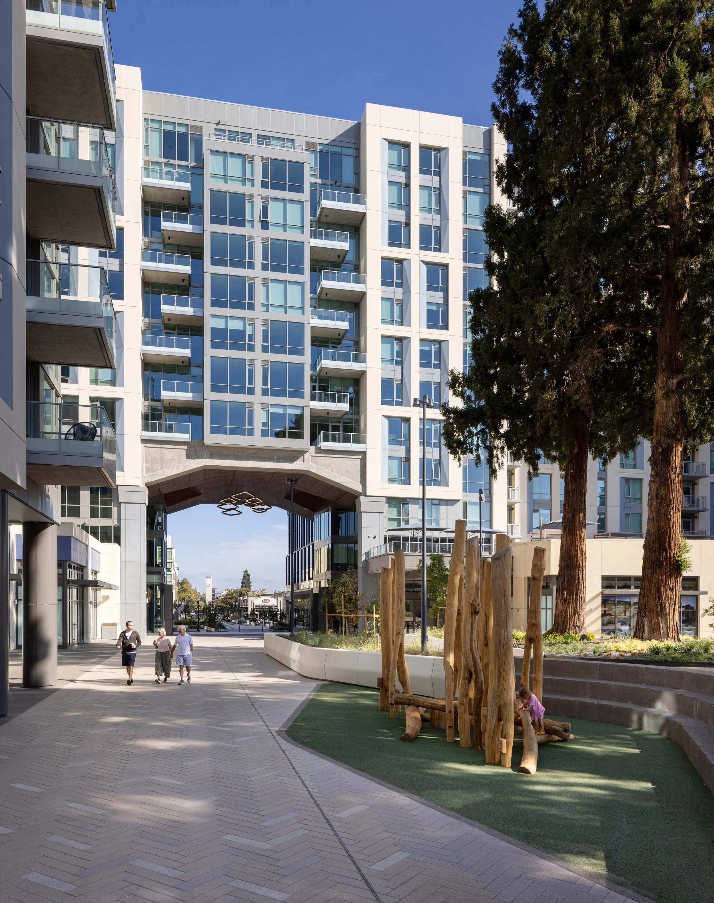 Modern residential building with glass balconies, a landscaped courtyard with large trees, wooden playground equipment, and people walking.