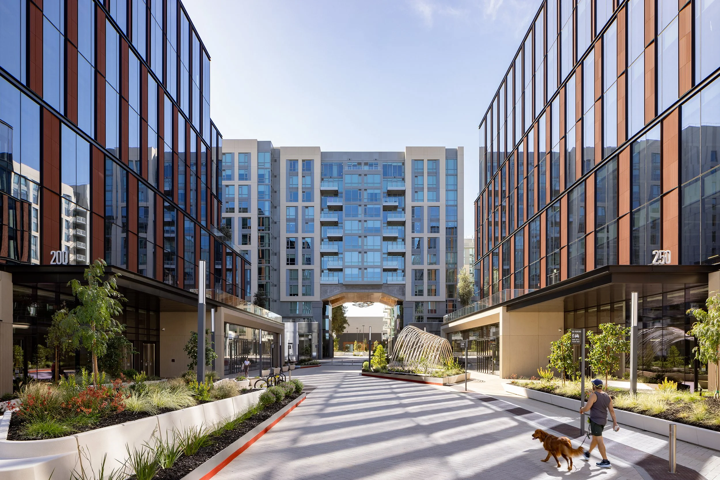 Modern apartment complex with two glass buildings on either side of a wide courtyard, featuring landscaping and a person walking a dog.