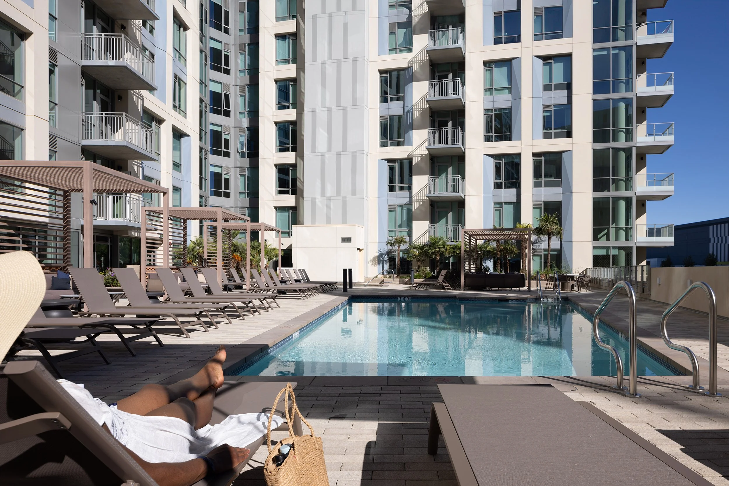 Person lounging sunbathing on poolside chair with bag nearby, view of swimming pool surrounded by lounge chairs and modern high-rise apartment buildings, clear blue sky.