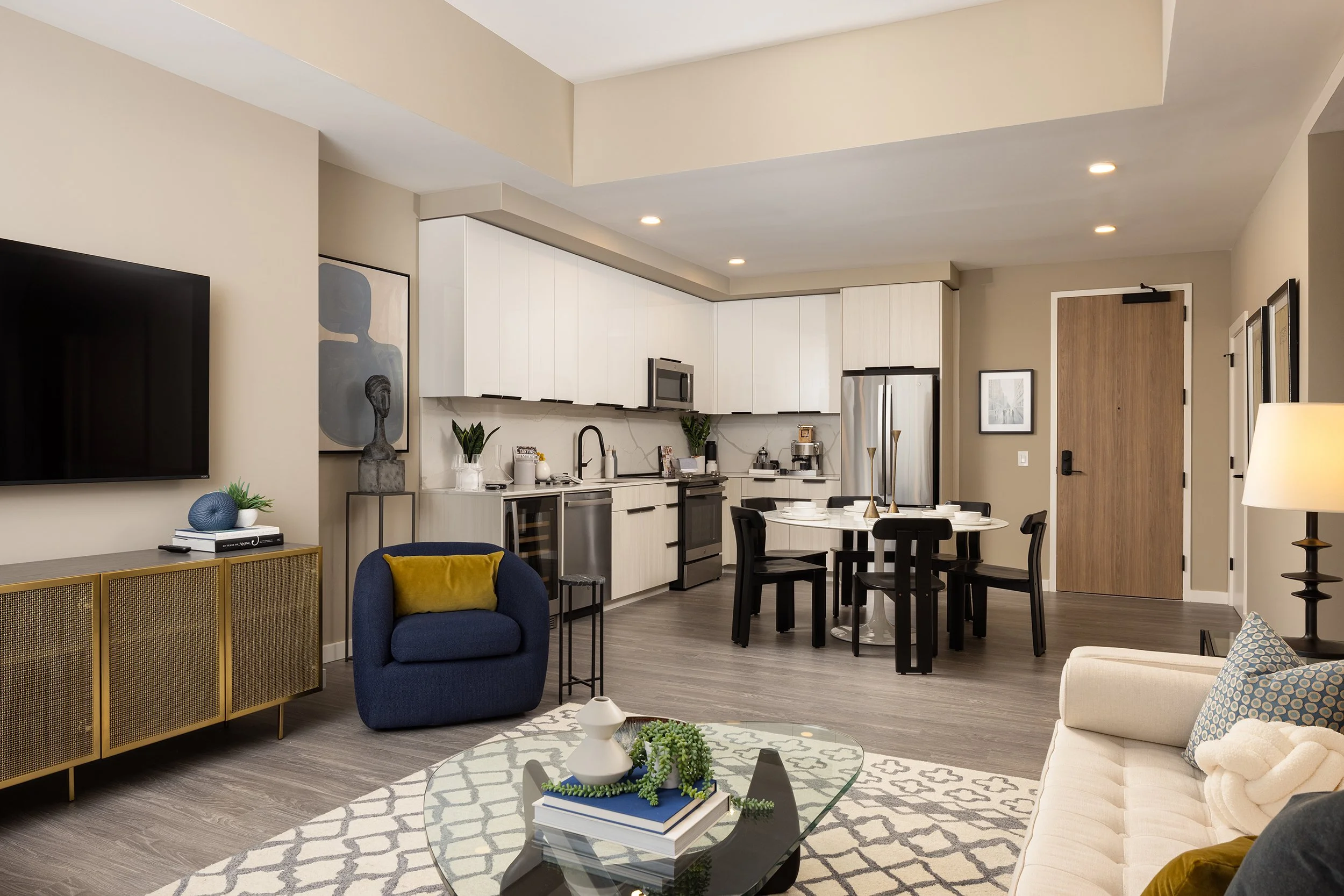 Open-concept living room and kitchen with modern decor, including a wall-mounted TV, a blue armchair, a white sofa with patterned pillows, a glass coffee table with decorative items, a round dining table with six black chairs, white cabinetry, stainl